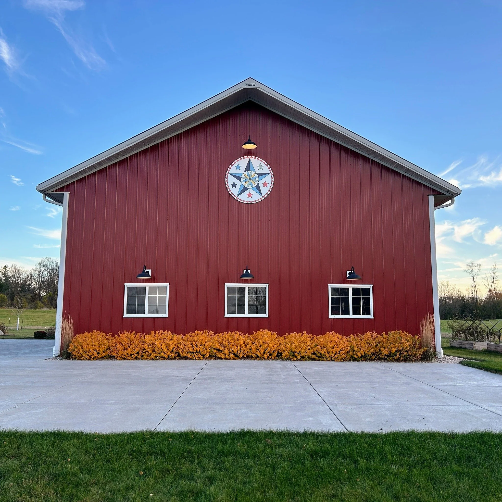 Red barn with three windows, black light fixtures, a circular star emblem near the roof, yellow flowers at the base, a sidewalk, green grass, and a blue sky with clouds.