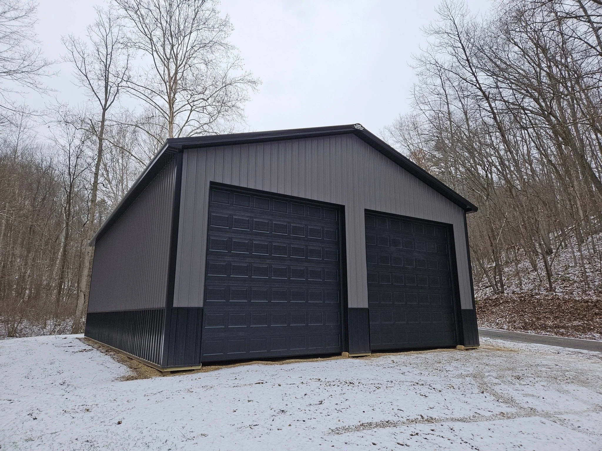 A large gray metal barn with two black garage doors situated on a snow-covered ground near a wooded area with leafless trees.