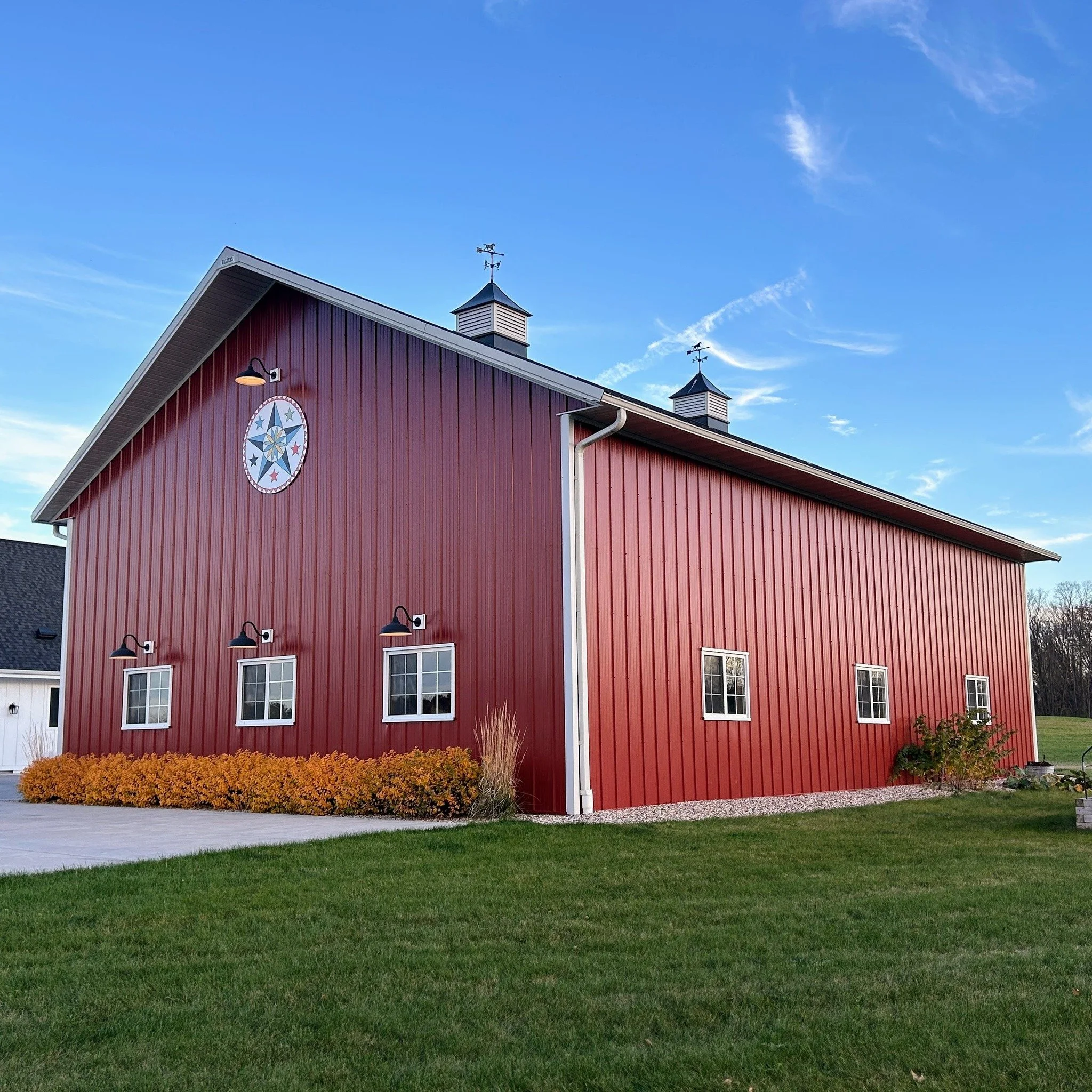 A large red barn with white-trimmed windows and black lamps, a decorative star and circle emblem on the front, set against a blue sky with some clouds, surrounded by green grass and some bushes.