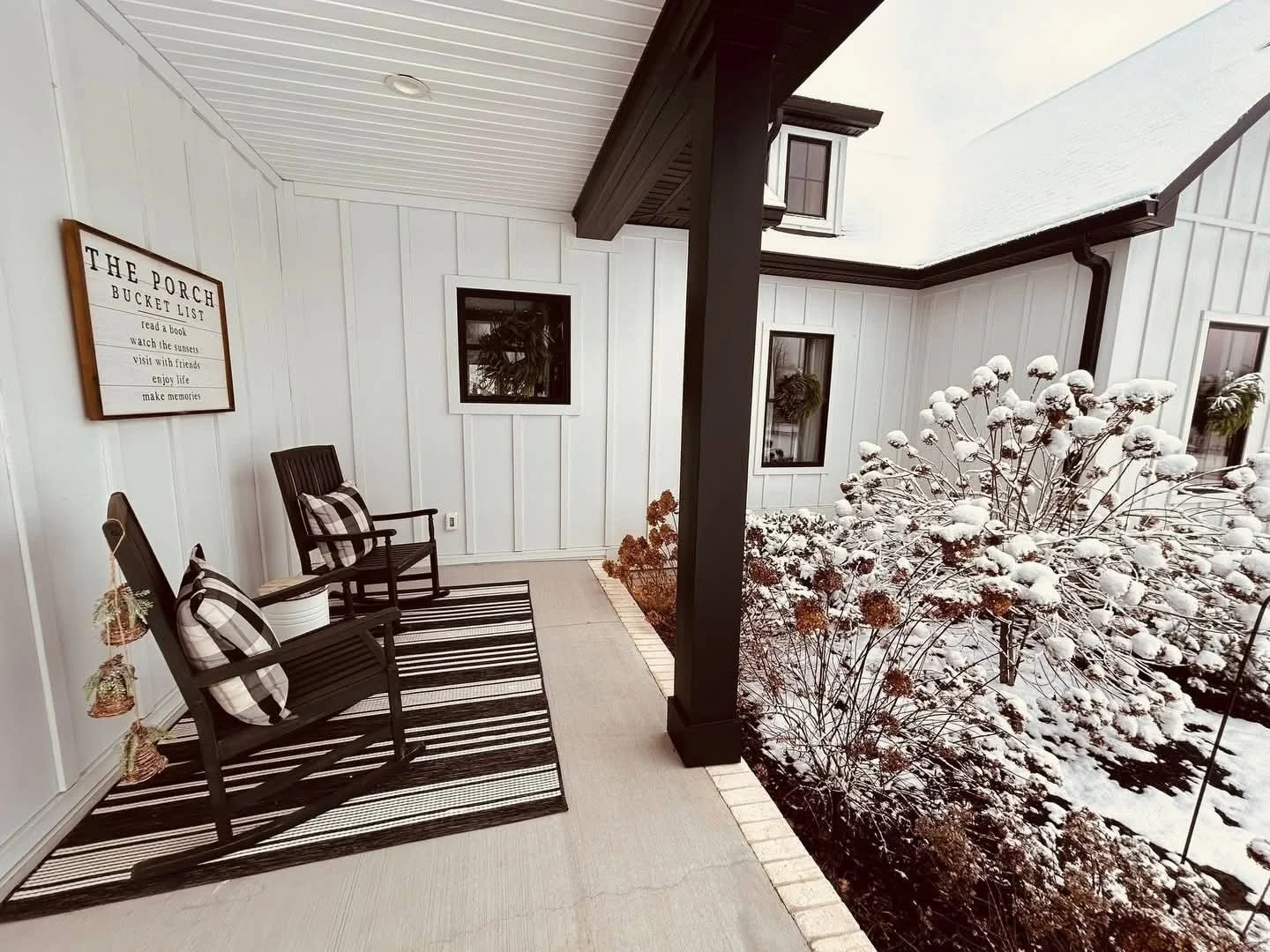 Covered porch with two black wooden chairs with plaid pillows, a striped black and white rug, a white wall with a sign, and snow-covered bushes outside in a winter setting.