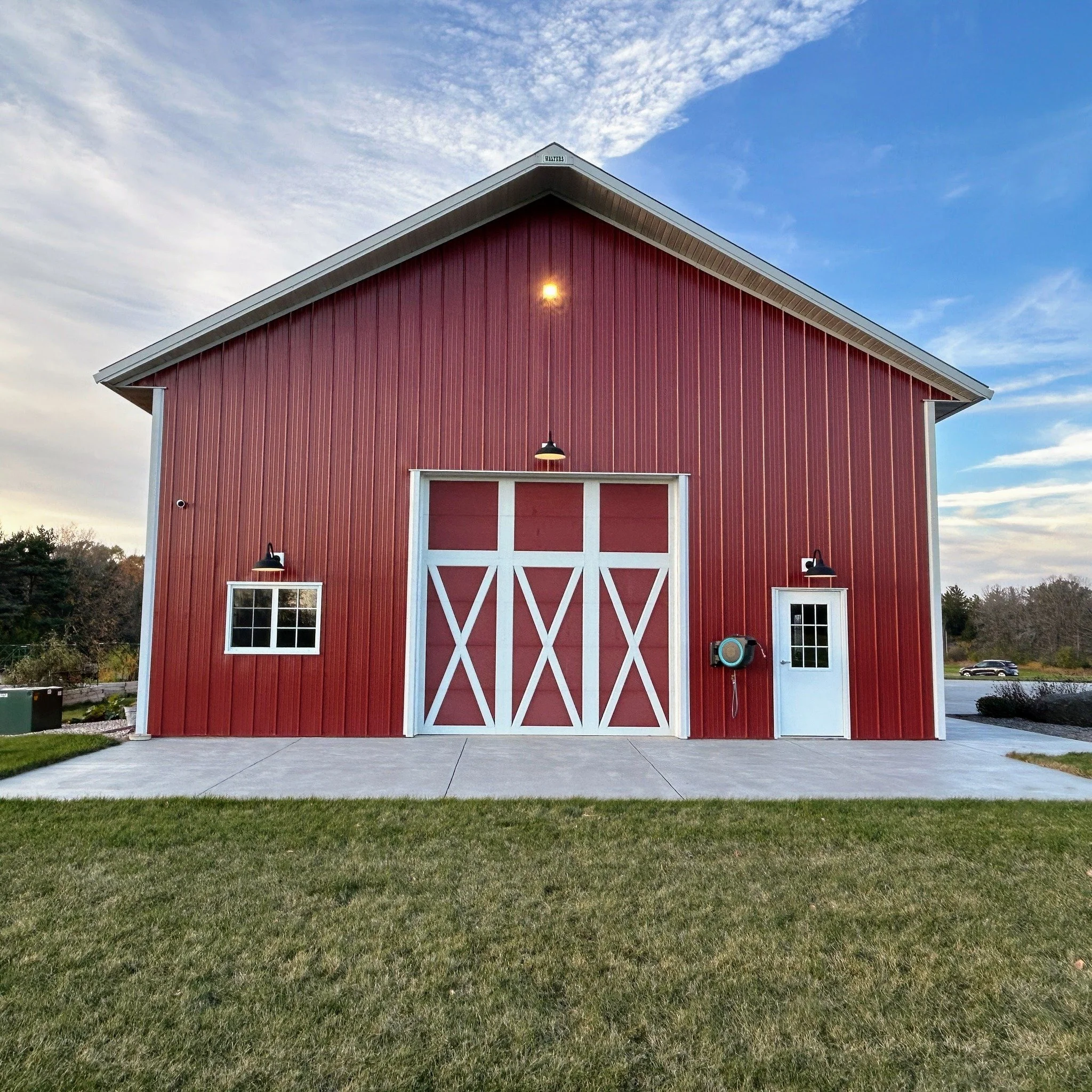 Red barn with white trim and large sliding doors, set on a concrete pad with green grass in the foreground, under partly cloudy sky.
