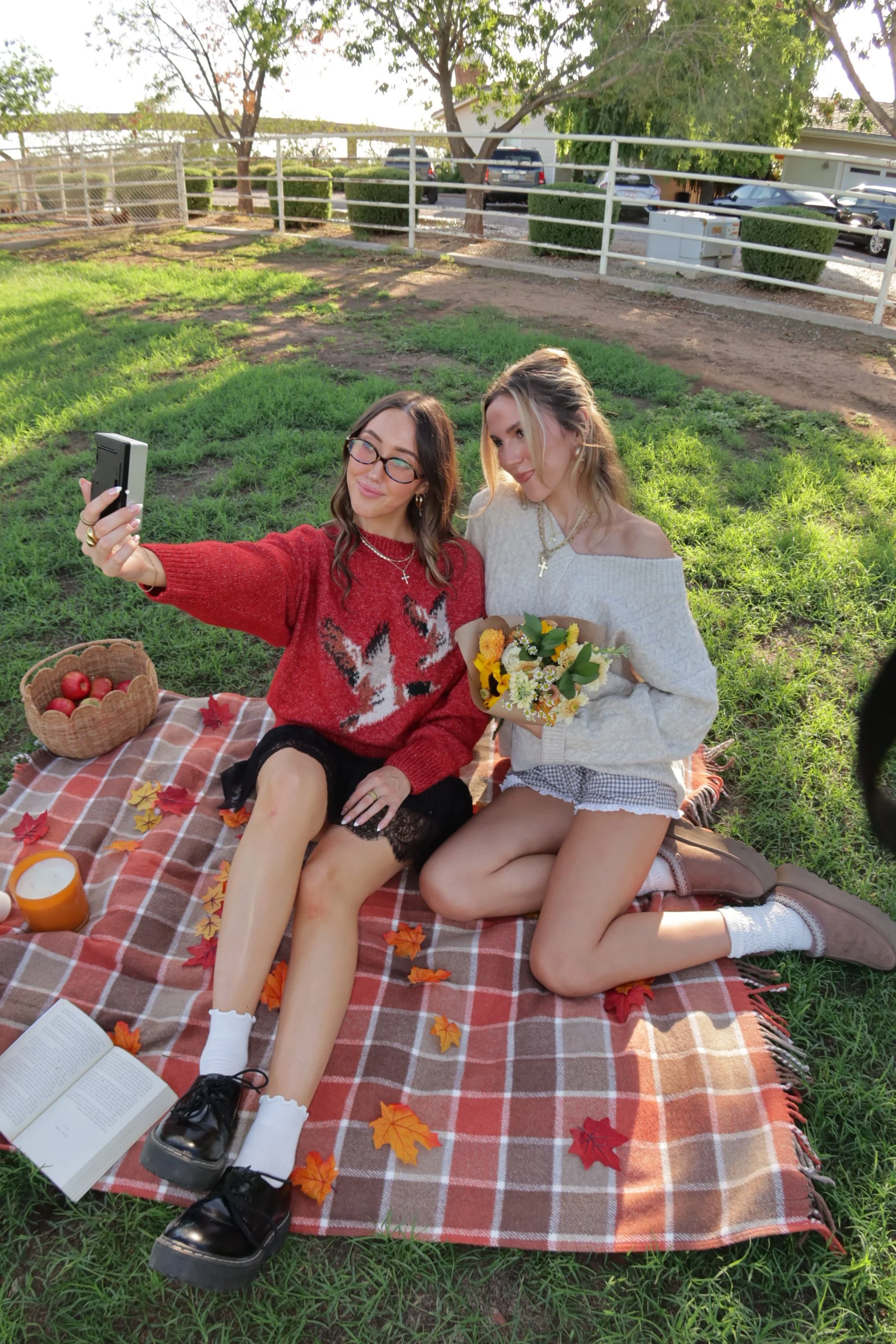 Two young women sitting on a picnic blanket outdoors, taking a selfie together. One woman holds a bouquet of flowers, and the other is smiling with glasses, wearing a red sweater with a fox pattern. There are autumn leaves, a book, a candle, and a basket of apples on the blanket.
