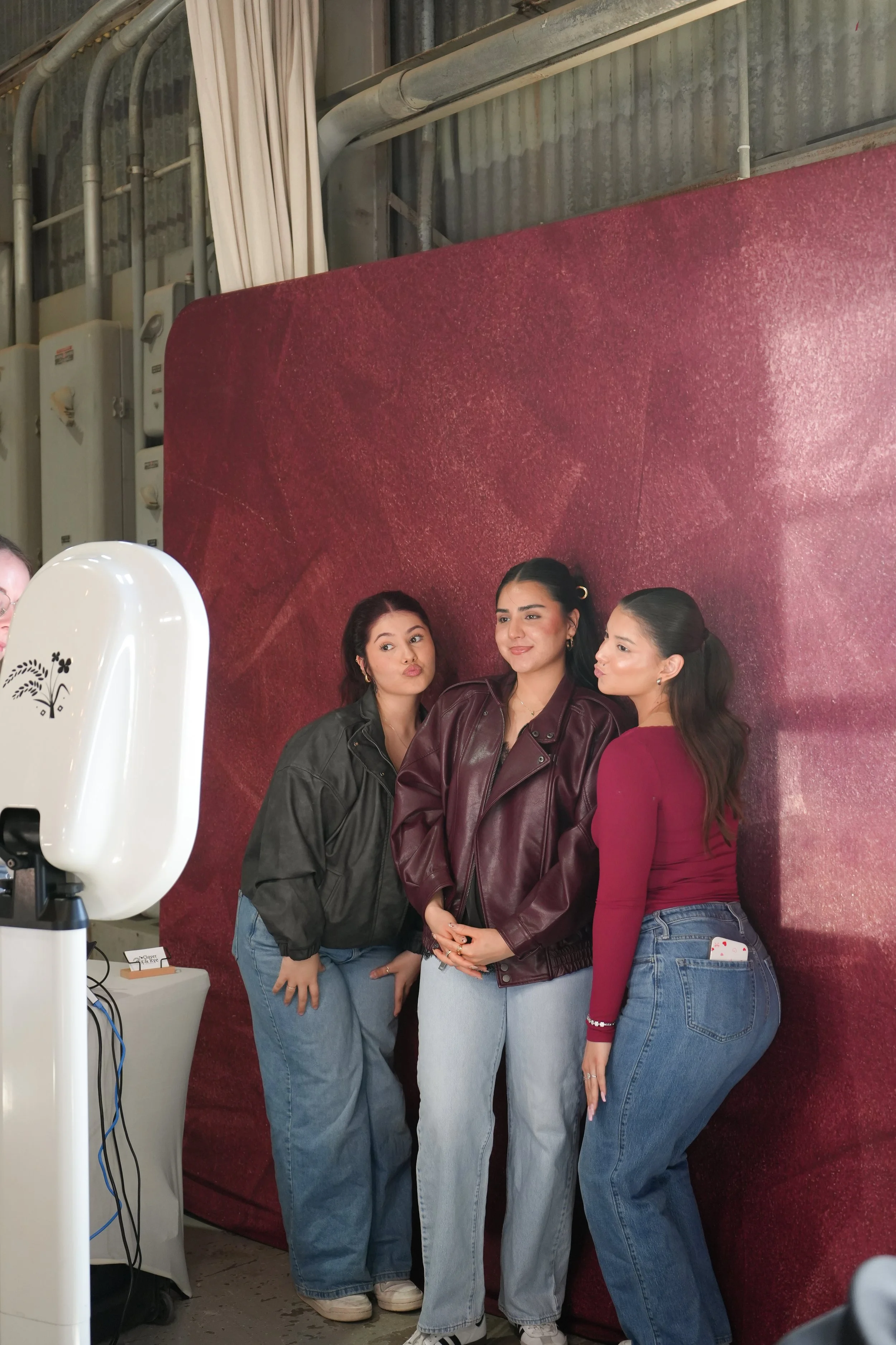 Three women posing for a photo, all with dark hair. The woman on the left is wearing a black jacket and blue jeans. The woman in the middle is wearing a red jacket and blue jeans. The woman on the right is wearing a red long sleeve top and blue jeans