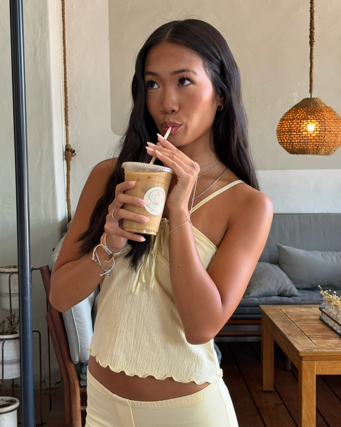 A woman with dark hair sipping iced coffee through a straw in a cafe setting.