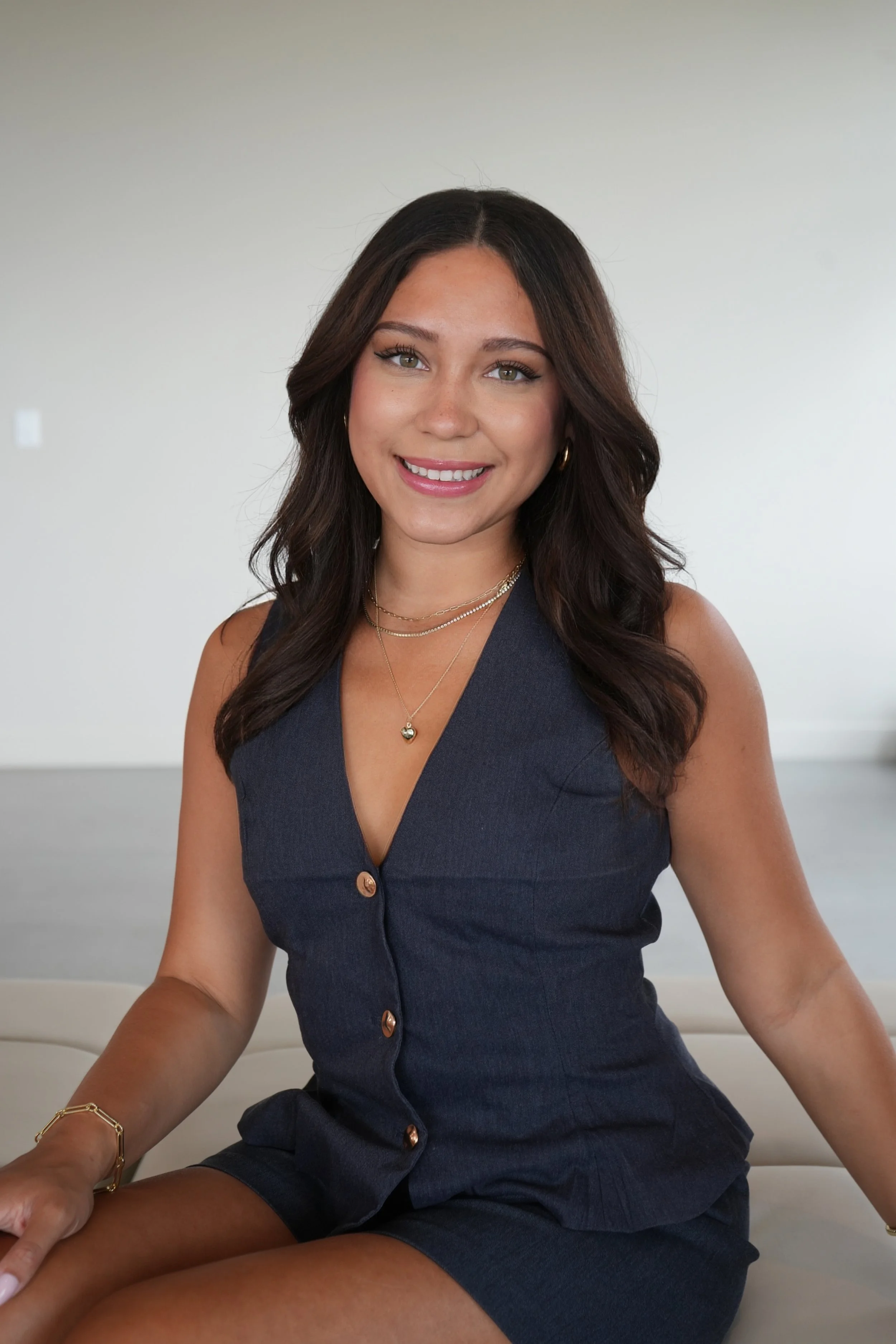 A woman with long dark hair, wearing a navy sleeveless dress, gold jewelry, and sitting on a white couch, smiling at the camera.