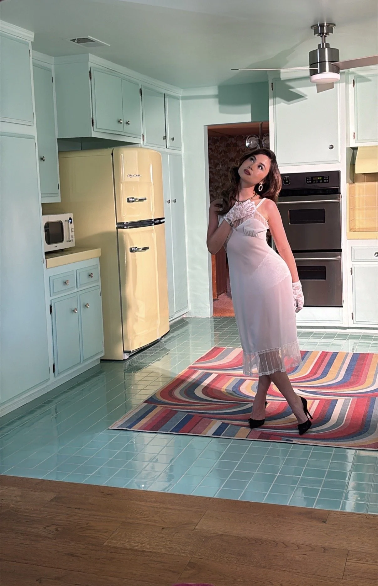 A woman in a white lace slip dress and black high heels posing in a retro-style kitchen with pastel blue cabinets, a yellow refrigerator, and a colorful striped rug.