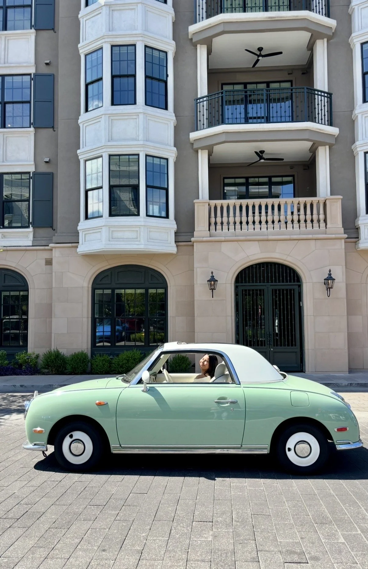 A woman sitting in a vintage mint green convertible car parked in front of a modern multi-story apartment building with large windows and balconies.