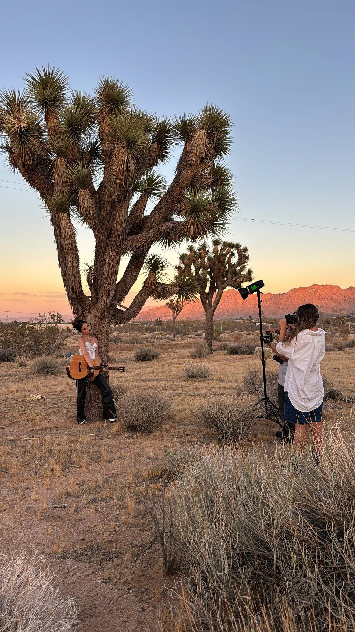 A photoshoot in the desert of a woman holding a guitar. There are two photographers holding cameras.