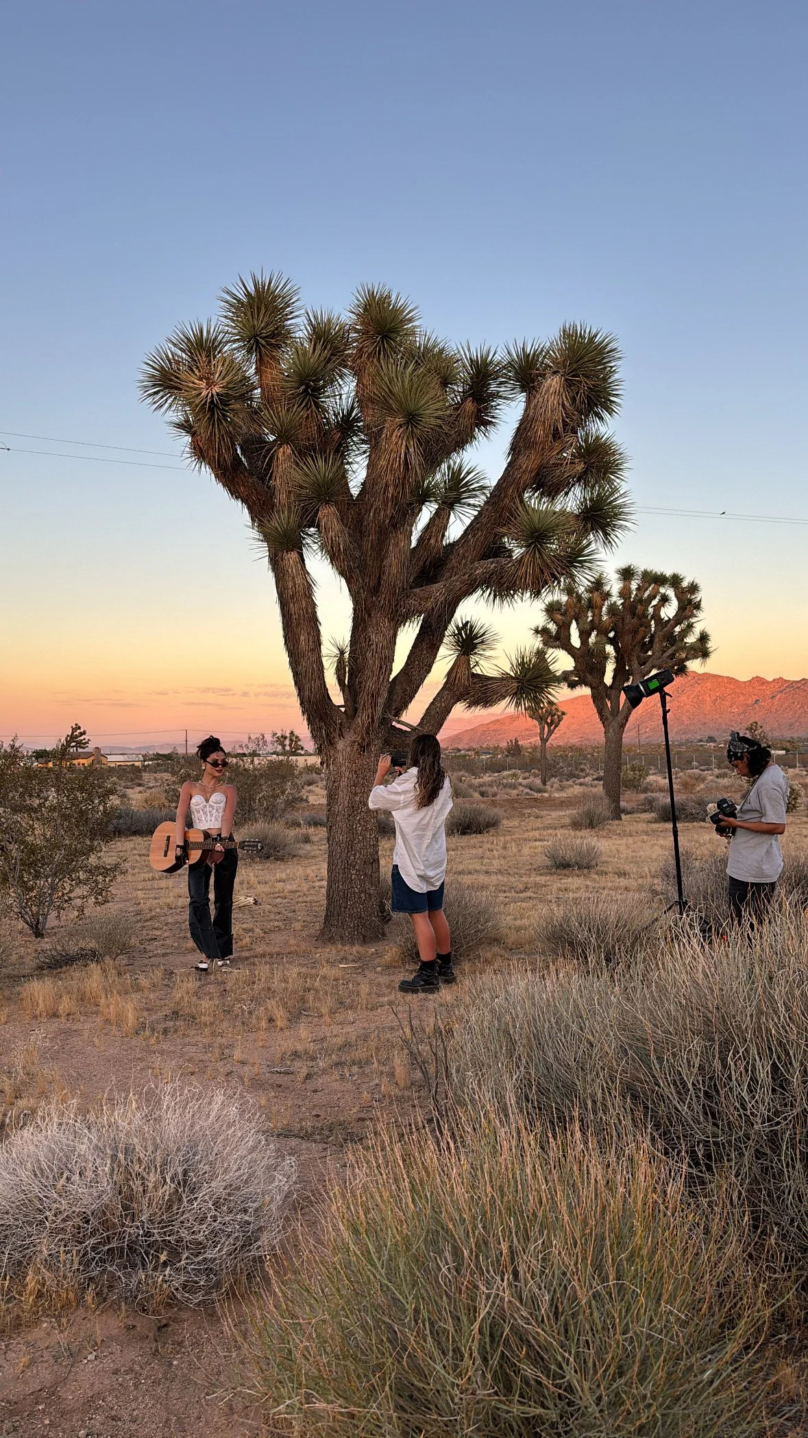 A desert photoshoot with a woman holding a guitar. There is a photographer wearing a white shirt and black pants.