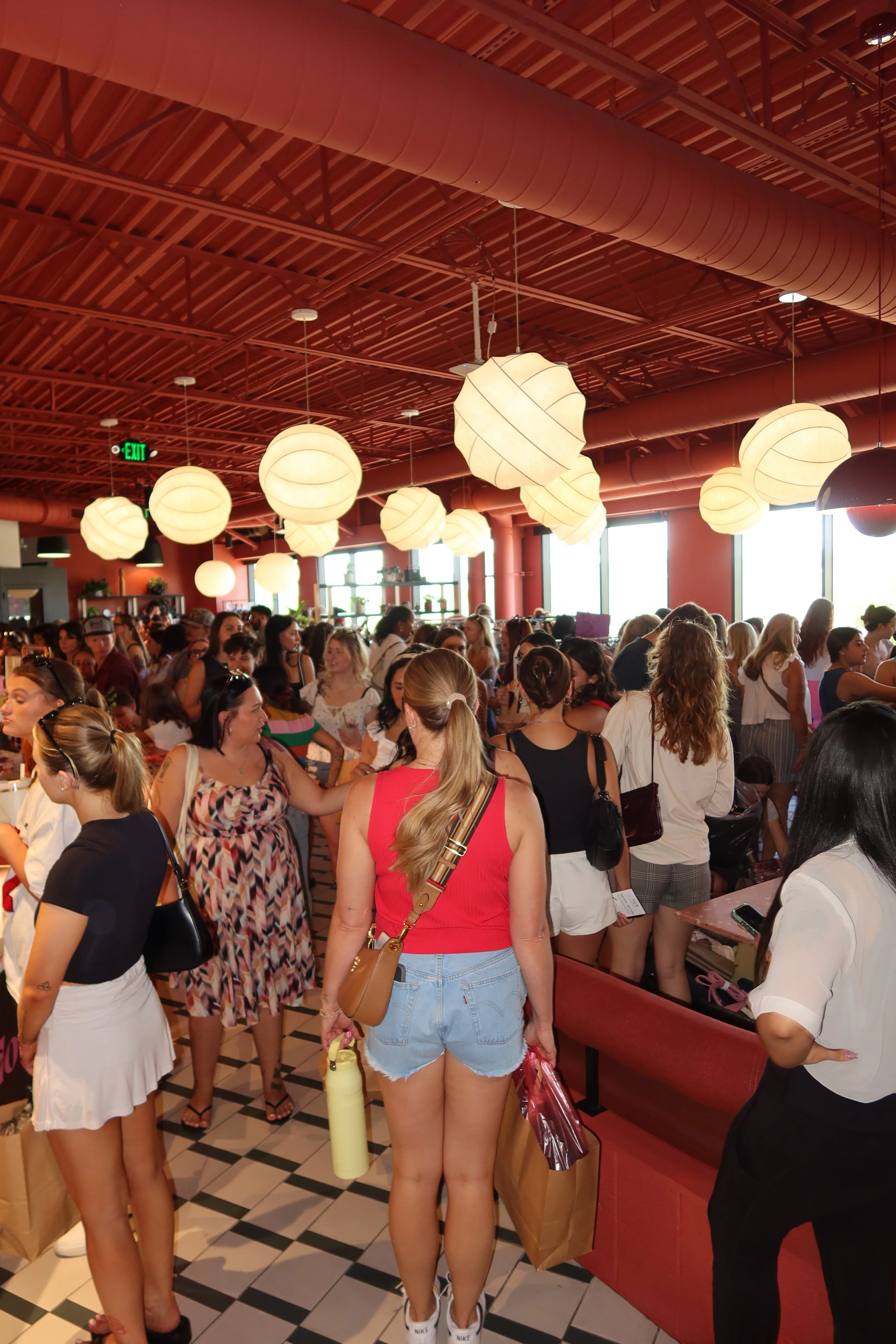 A thrift market scene with a room full of women. The focal point is a blonde girl wearing a red tank top and blue jean shorts. She is holding a yellow water bottle and a brown bag.