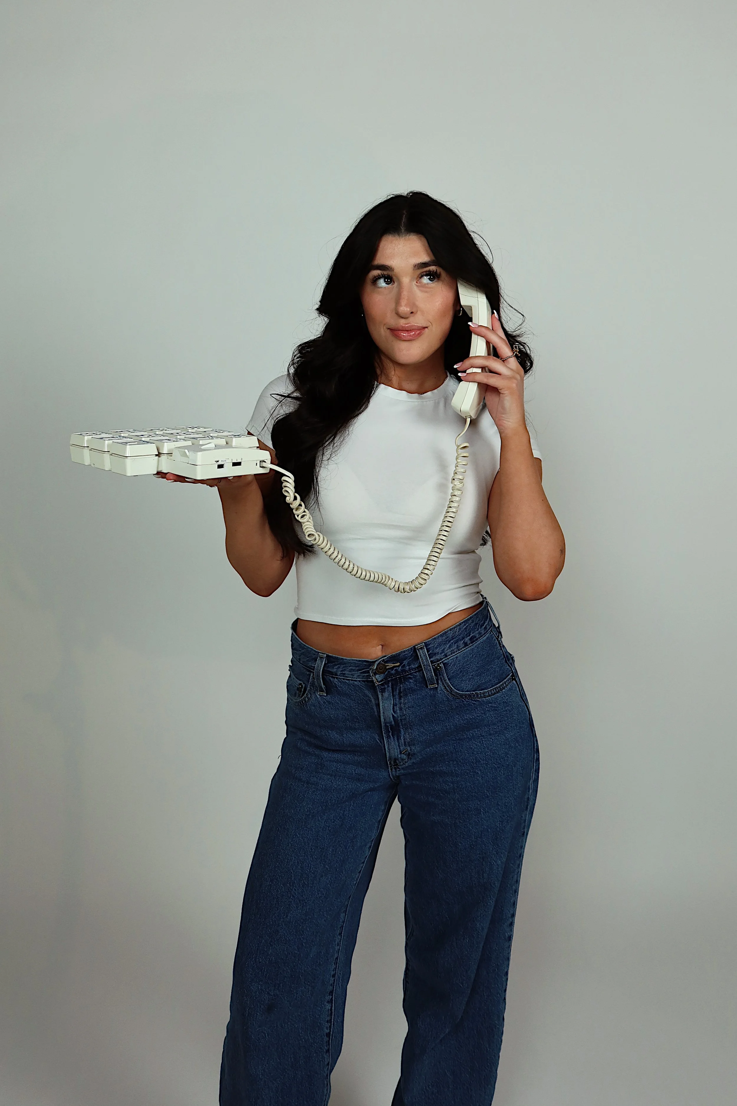 The head of creative and growth for Going Social Club, Kayla. She is posing with a white landline phone. She is wearing a white t-shirt and blue jeans.