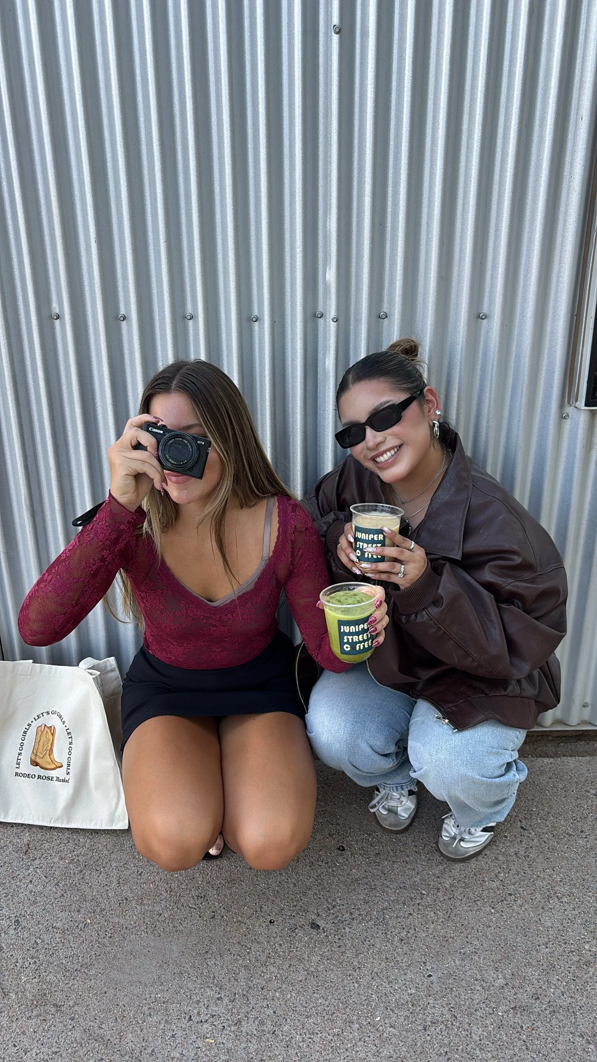 Two women holding a coffee and matcha from Juniper Street Coffee. The woman on the left is wearing a red long sleeve top and black skirt. She is holding a black camera. The woman on the right is wearing a brown jacket and blue jeans.
