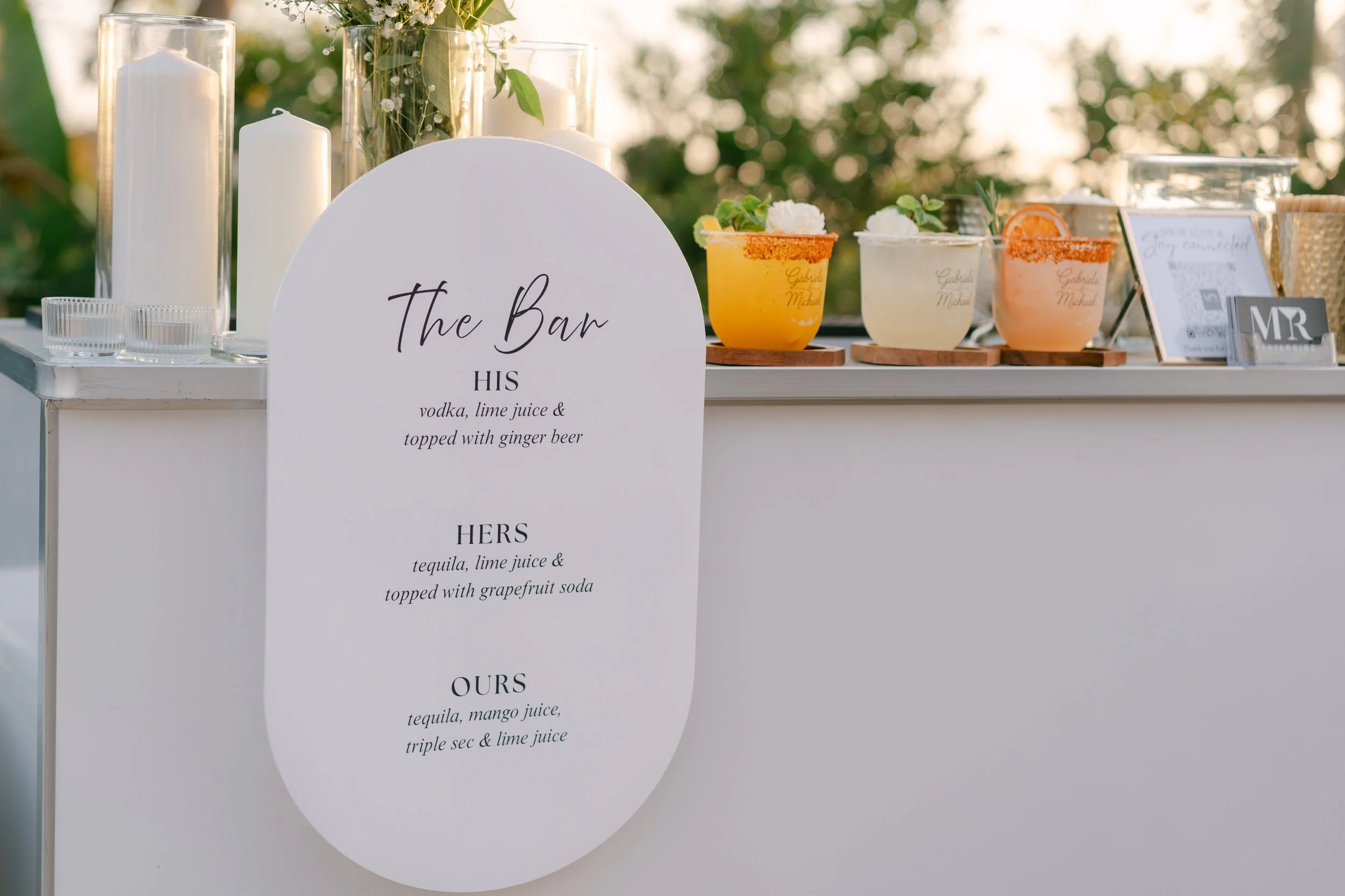 A white bar with a menu sign listing three cocktails: one with vodka, lime juice, and ginger beer; another with tequila, lime juice, and grapefruit soda; and a third with tequila, mango juice, triple sec, and lime juice. Behind the bar are candles and colorful cocktails garnished with fruit slices and herbs.