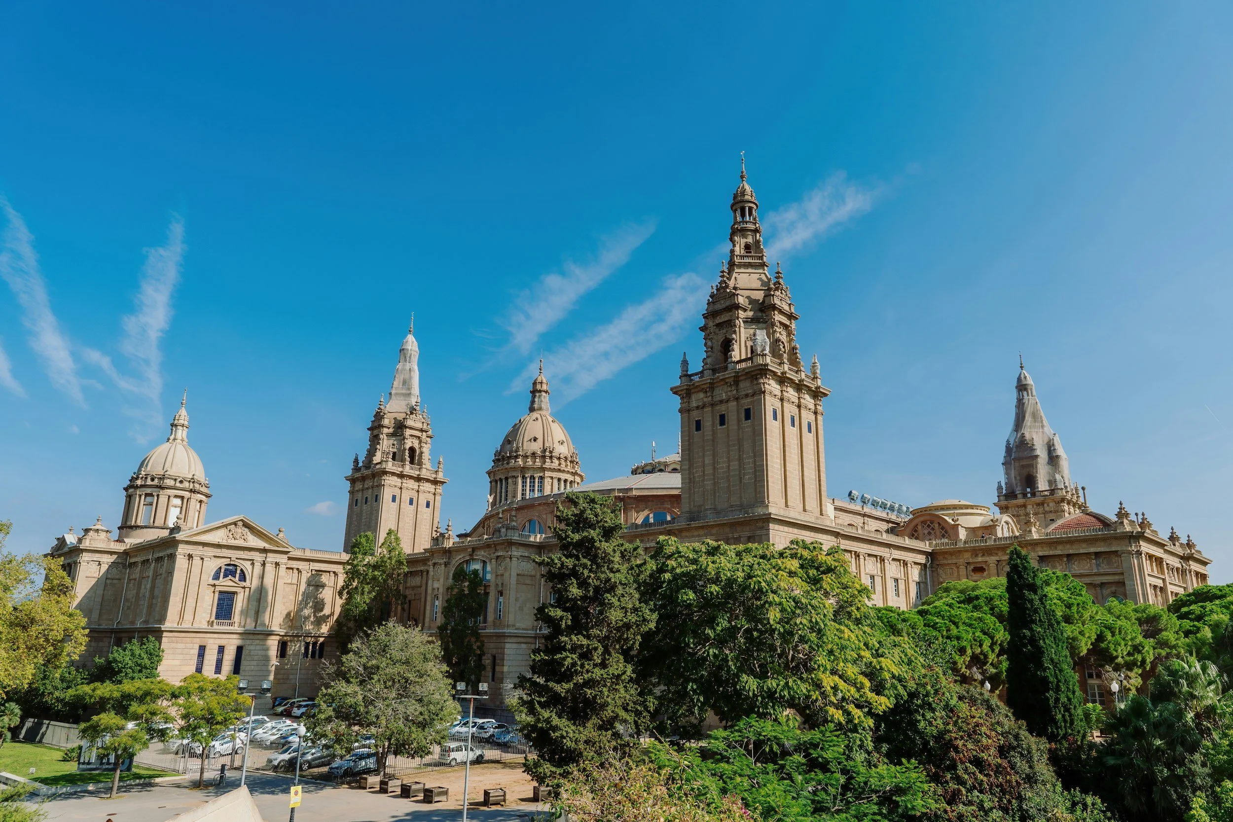 A historic building with multiple towers and domes, surrounded by lush green trees and parking lot, under a clear blue sky.