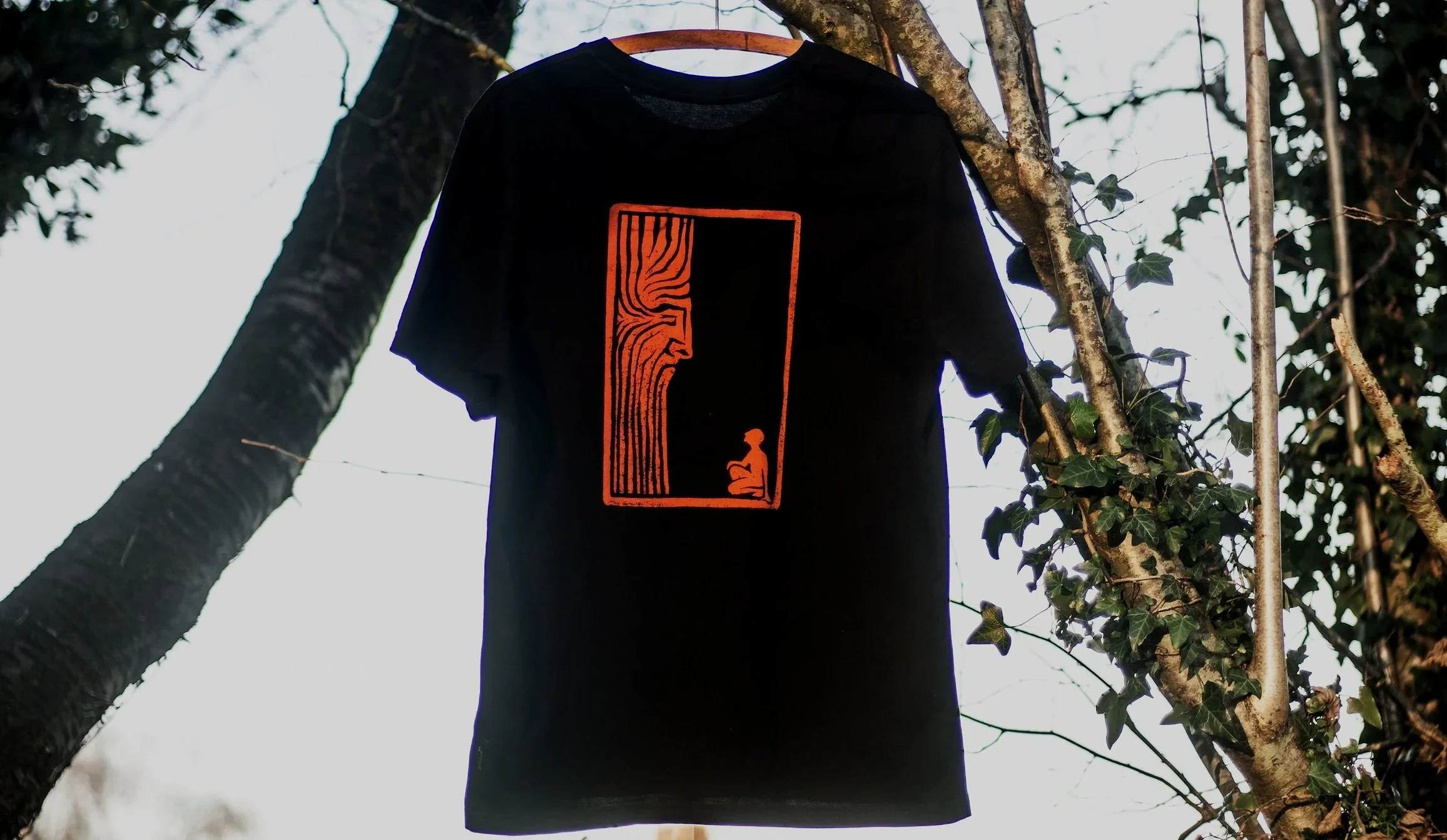 Black T-shirt hanging outdoors on a tree branch, featuring a red graphic design of person kneeling in front of Old One spirit design printed from woodblock print, surrounded by leaves and branches.