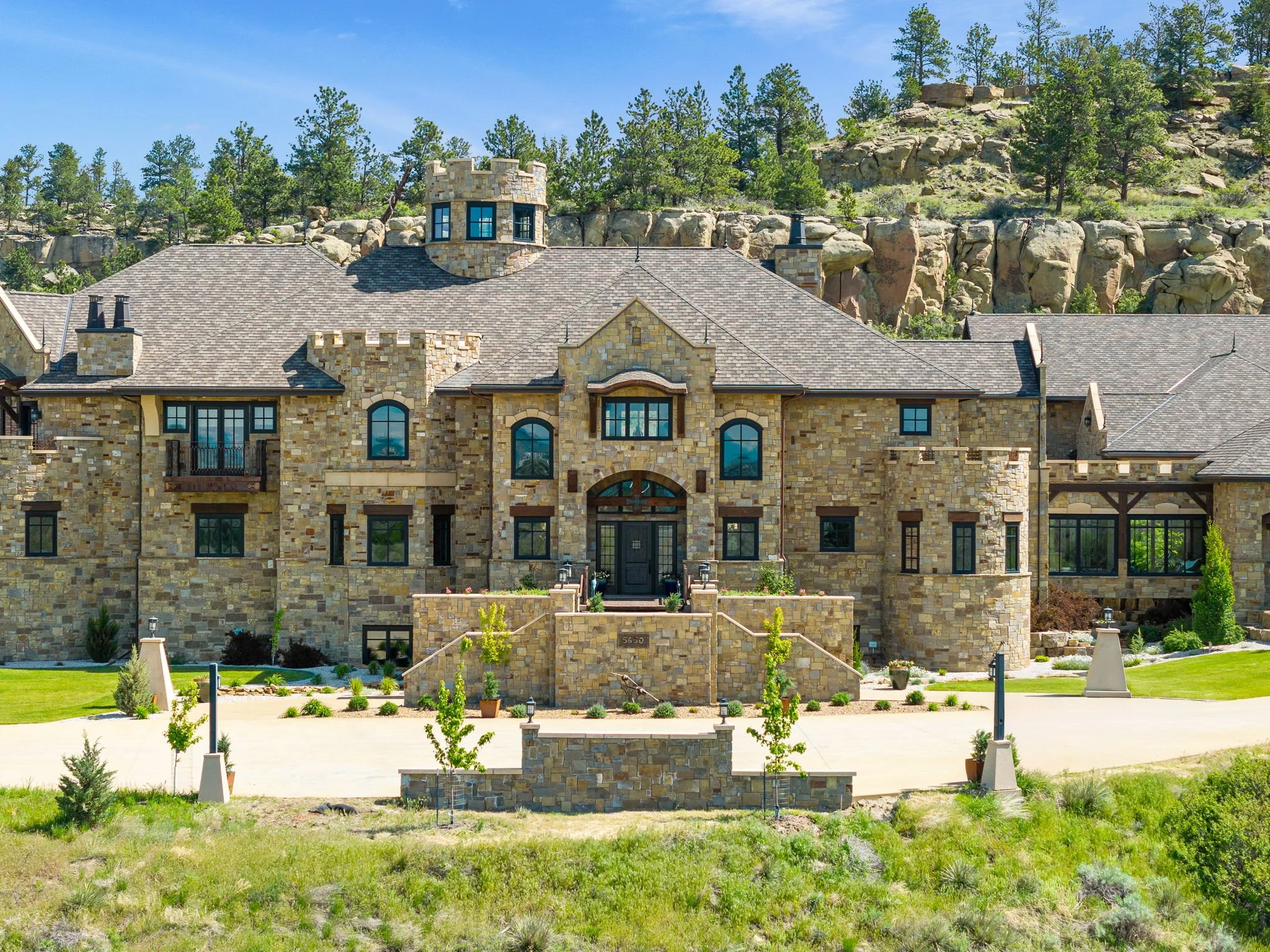 A large, luxurious stone mansion with multiple levels, small turrets, and numerous windows, surrounded by a manicured lawn and landscaping, situated at the base of rocky hills with trees.