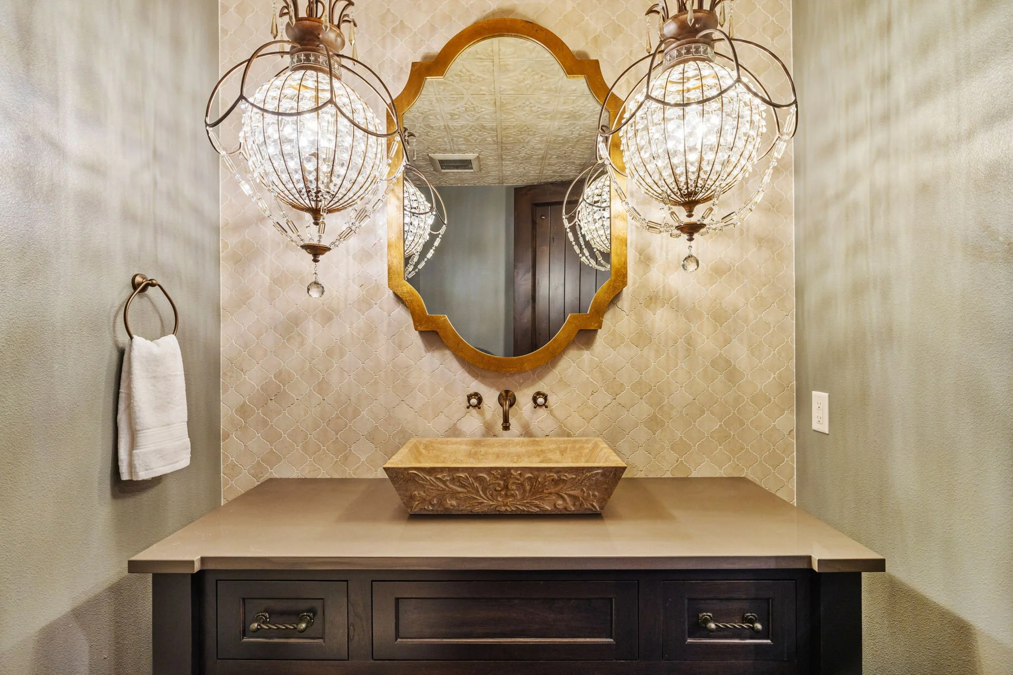 Elegant bathroom vanity with a carved stone sink, a large gold-framed mirror, and ornate hanging chandeliers, with a towel on a ring on the left wall.