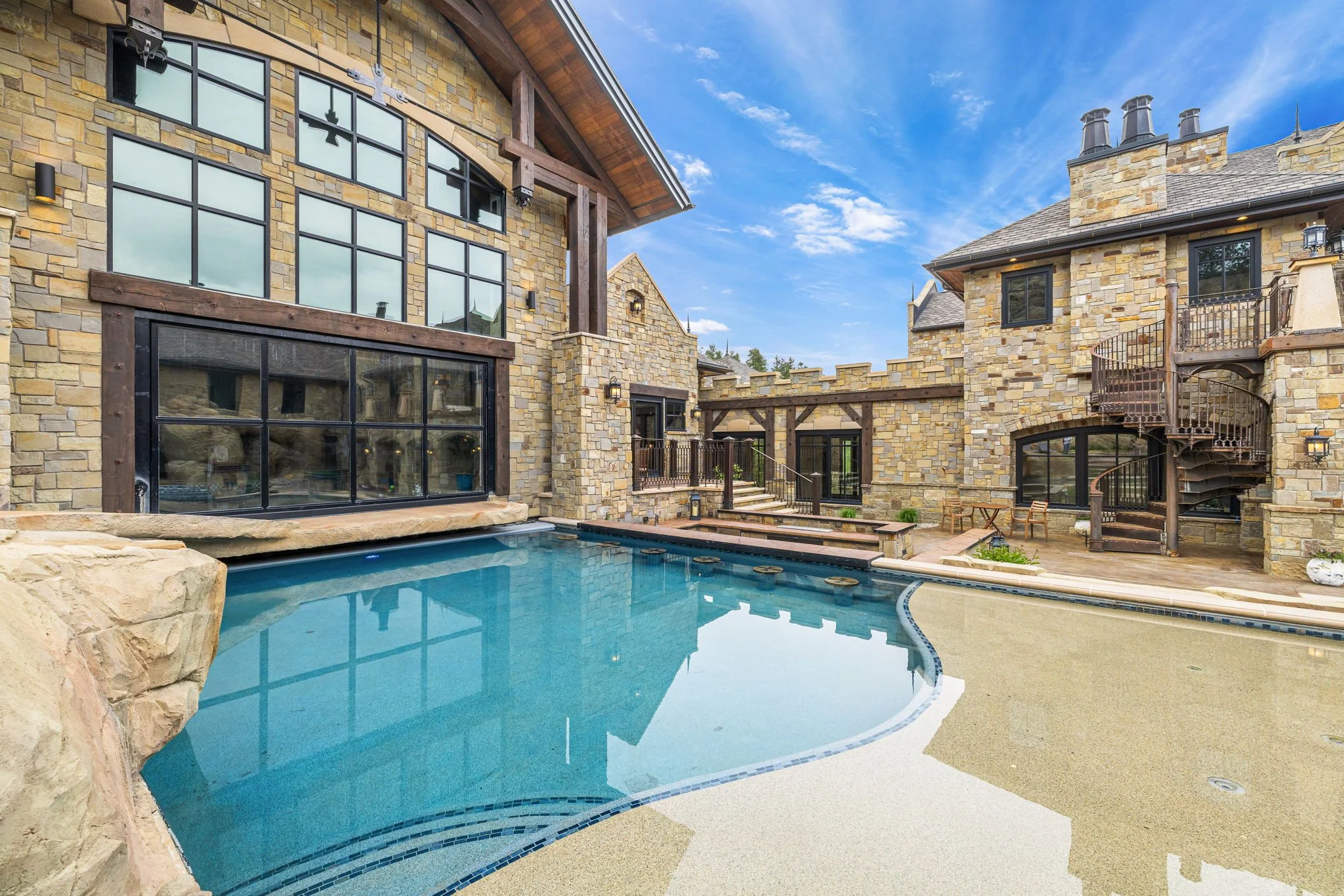 A backyard view of a stone house with large windows and a swimming pool, featuring a patio with chairs and outdoor spiral staircase.