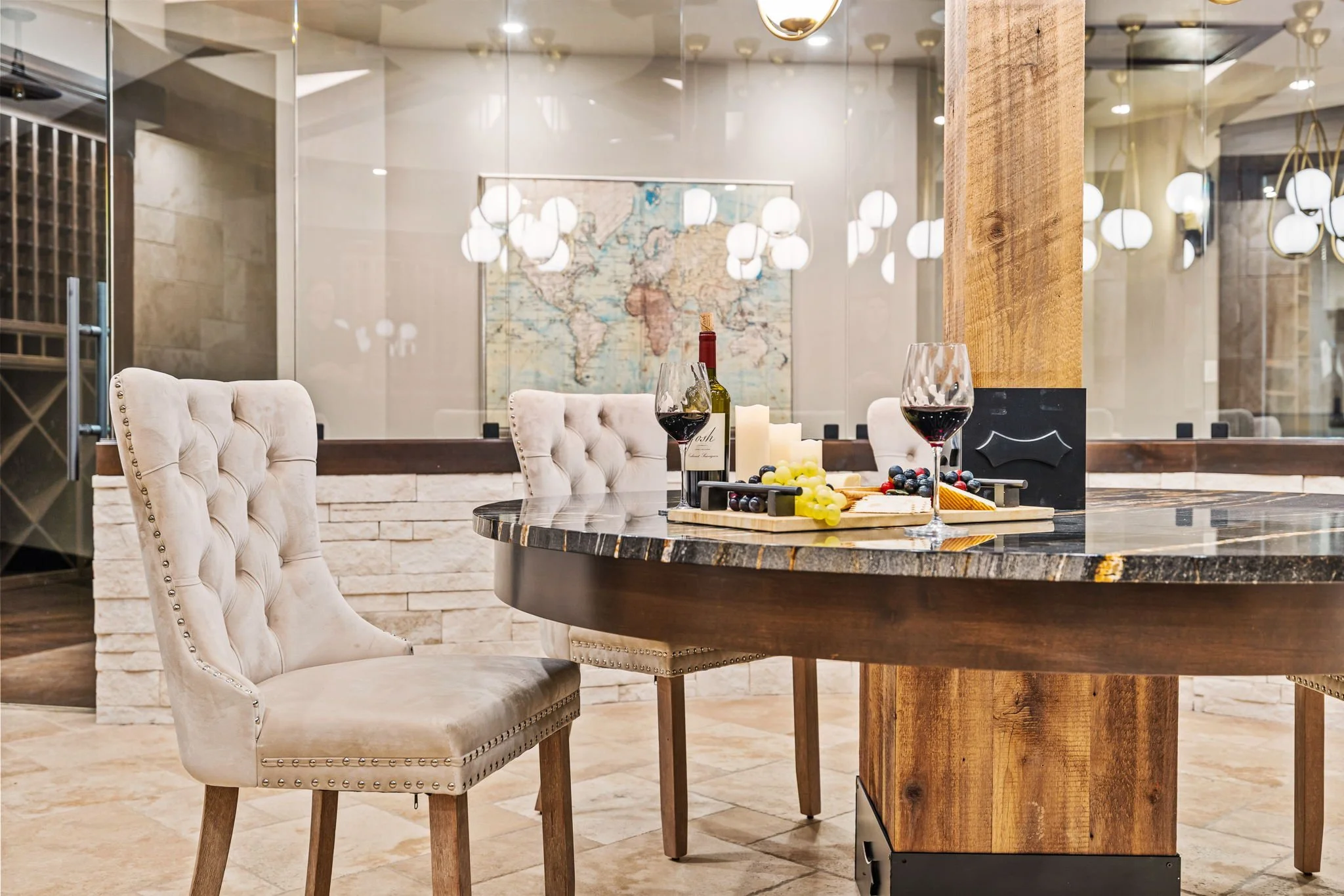 A dining area with a round marble-topped table set with wine glasses, a wine bottle, and a cheese platter, surrounded by beige upholstered chairs, with a world map on the wall behind and a glass divider reflecting modern hanging lights.
