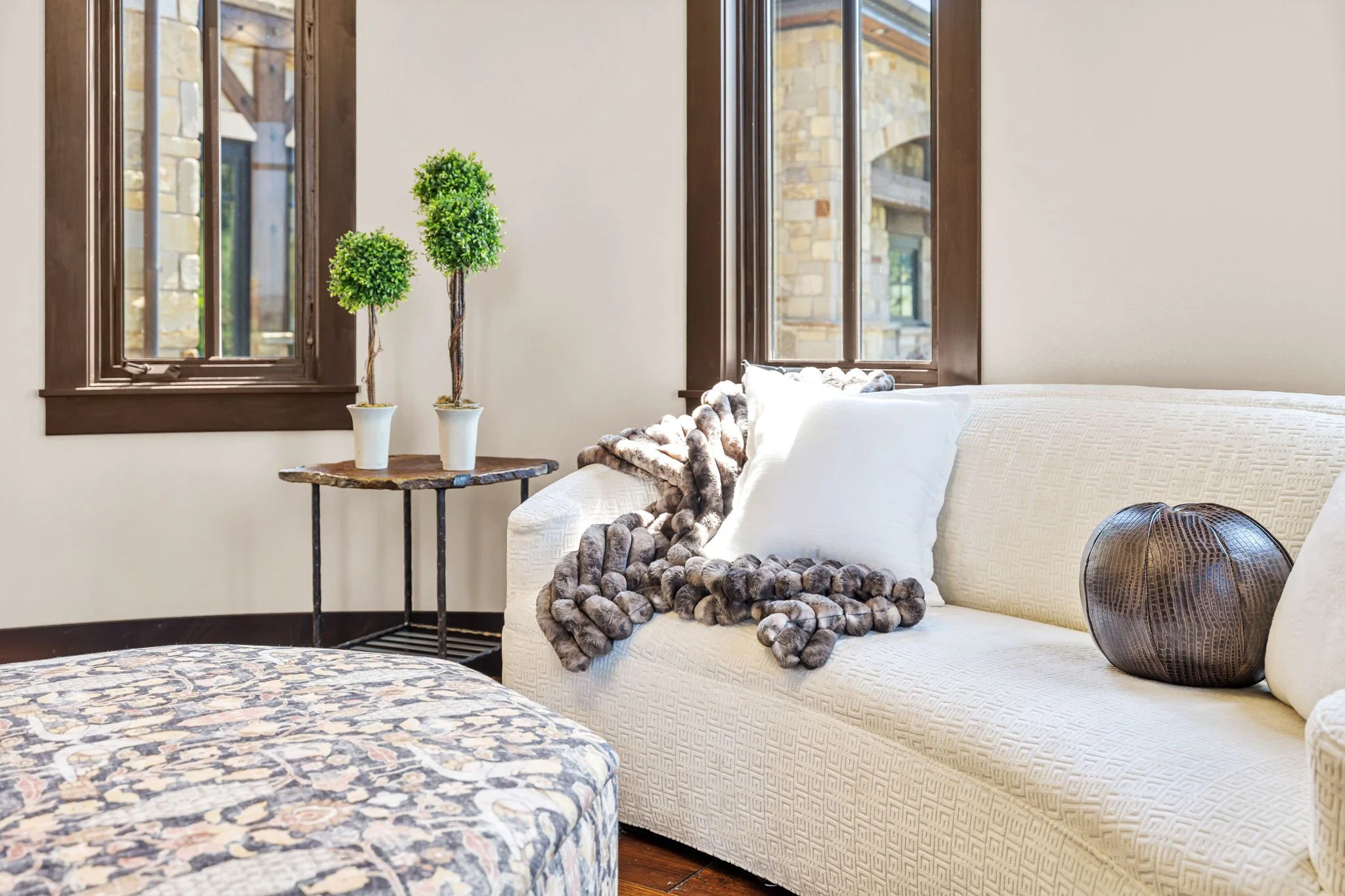Bright living room with a cream-colored sofa, two white pillows, a fur throw, a round decorative brown pillow, and a small table with two potted topiary plants, large windows with dark brown frames, and stone walls outside.