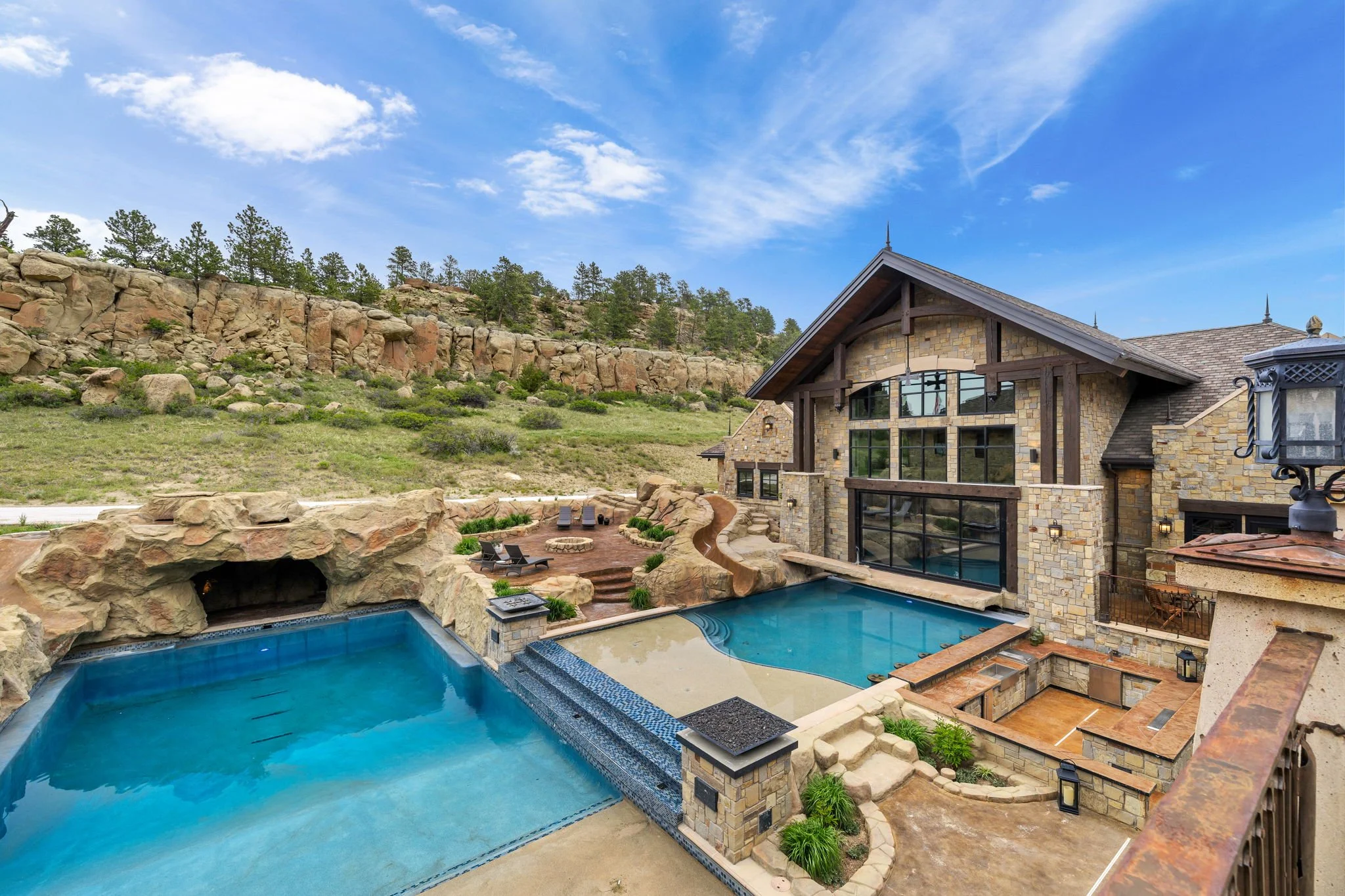 Luxury house with stone exterior, large windows, pool with waterfall, outdoor seating, and scenic rocky hillside with trees under a partly cloudy sky.