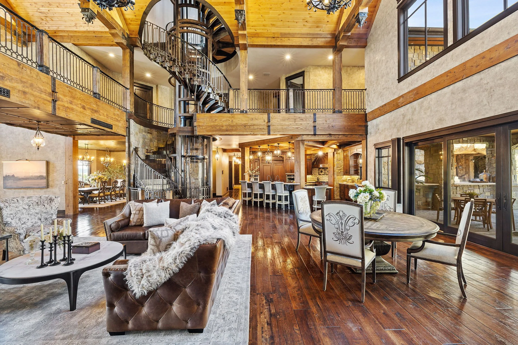 Spacious living room with high wooden ceilings, a central spiral staircase, large windows, and contemporary furniture including a leather sofa with fur throw, a dining table, and kitchen area in the background.