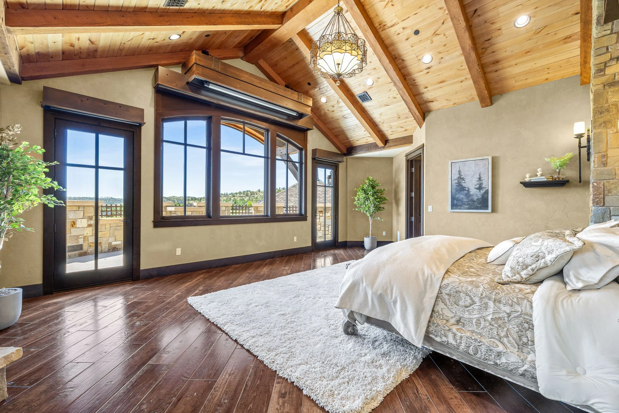 A spacious bedroom with a wooden vaulted ceiling, large windows with black trim, and a sliding glass door leading to a balcony. The room features a bed with white and beige linens, a white shag rug, potted plants, and wall decor. Recessed lighting an