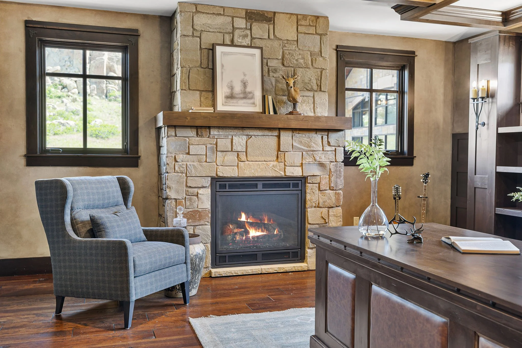 A cozy living room featuring a stone fireplace with a mounted deer head and framed artwork, a plaid armchair with a pillow, a wooden table with decorations, and two windows with black trim showing outdoor scenery.