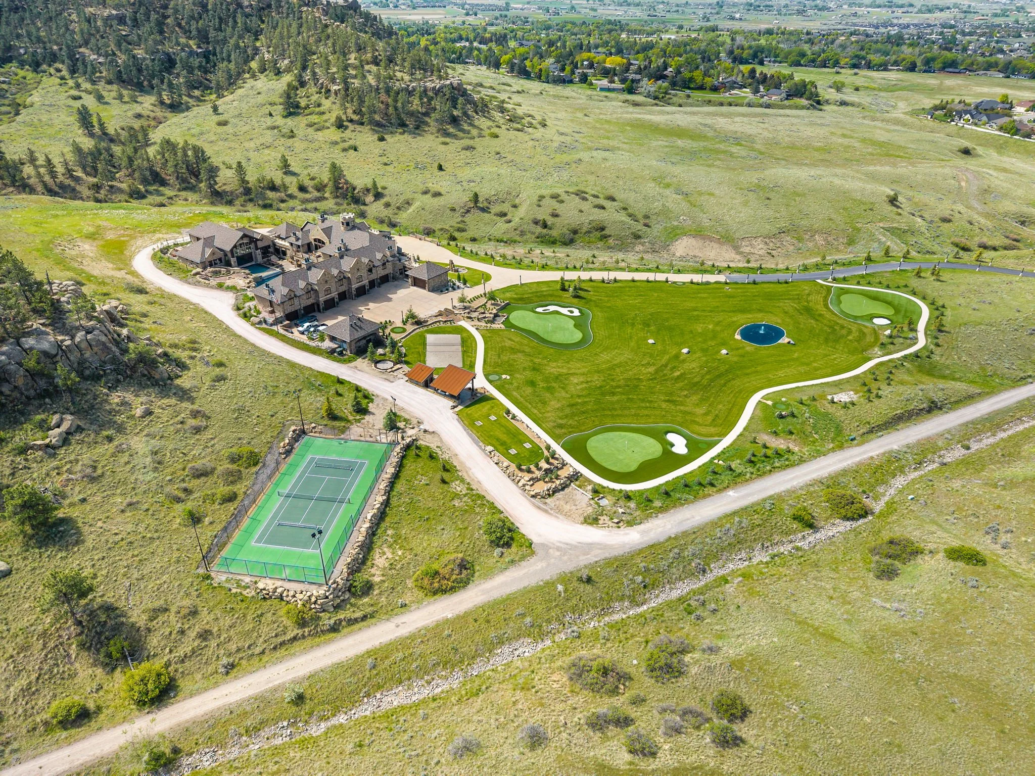A large estate with a mansion, tennis court, and a miniature golf course, surrounded by open grassy land and distant city views.