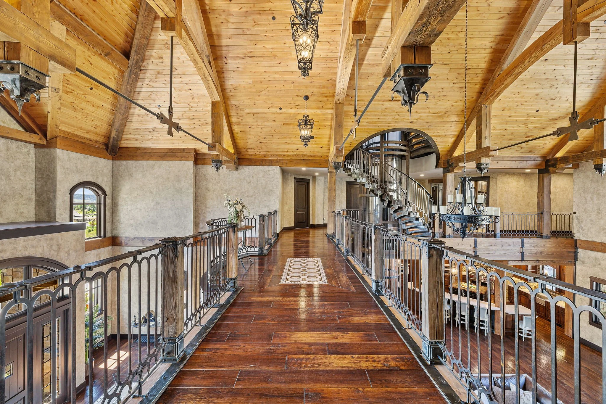 Interior view of a house with a high wooden ceiling, a narrow wooden walkway with decorative metal railings, a spiral staircase, and chandeliers.