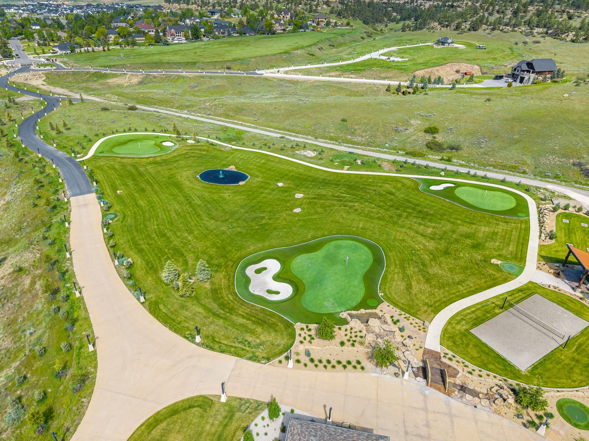 Aerial view of a golf course with three holes, a pond, and surrounding homes and roads in a hilly landscape.