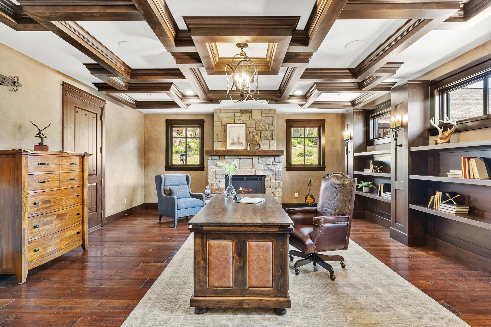 A spacious office with a large wooden desk, two armchairs, a stone fireplace, wooden ceiling beams, dark wood bookshelves, and windows showing greenery outside.