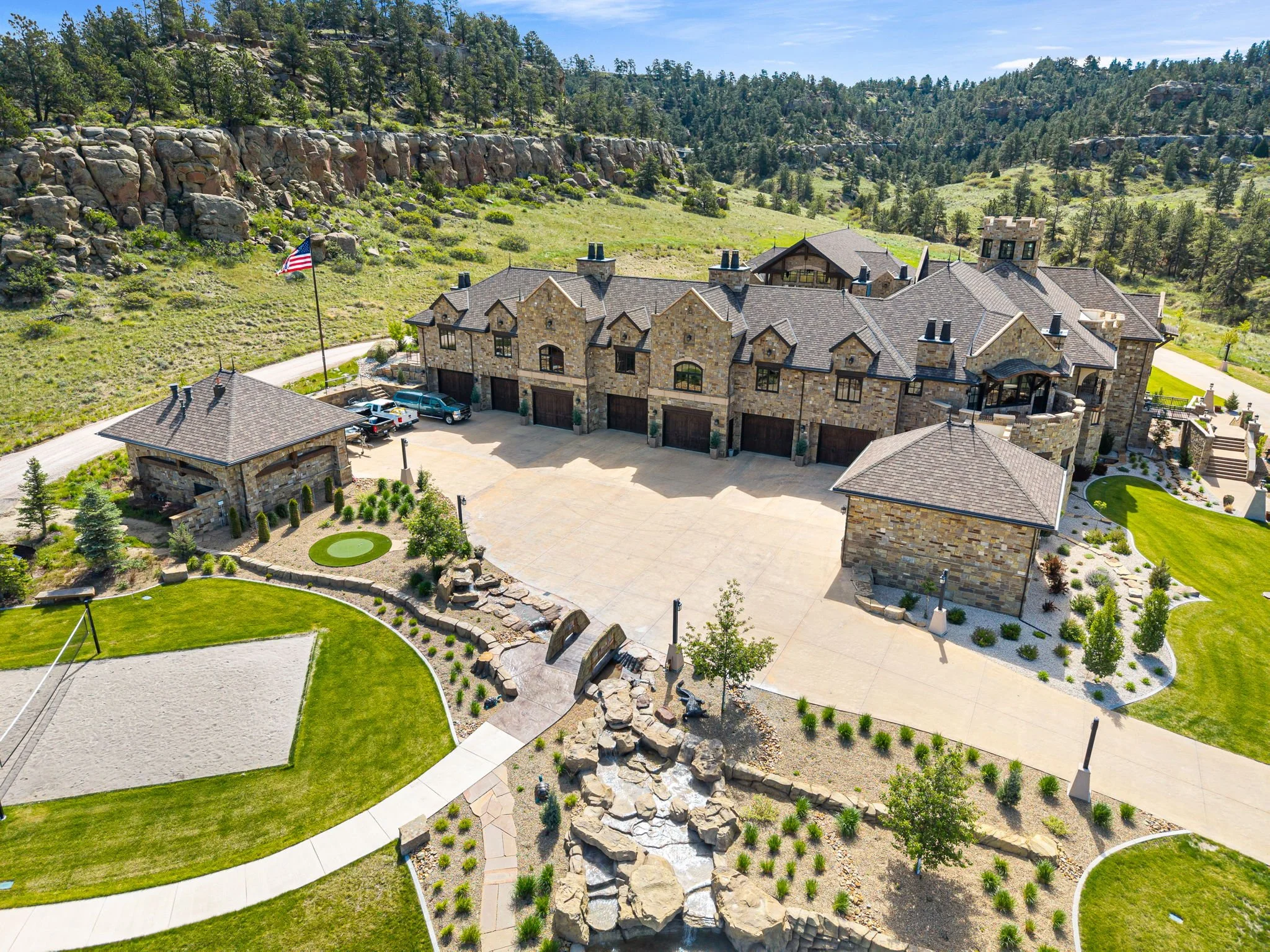 A large stone mansion with multiple garages, a paved driveway, landscaped yard, and water feature surrounded by trees and rocky hills.