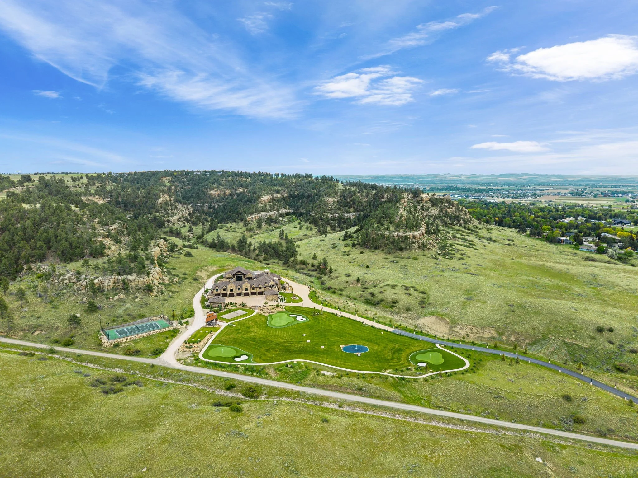 A large house with a golf course and tennis court on a grassy hillside, surrounded by trees and mountains under a partly cloudy sky.