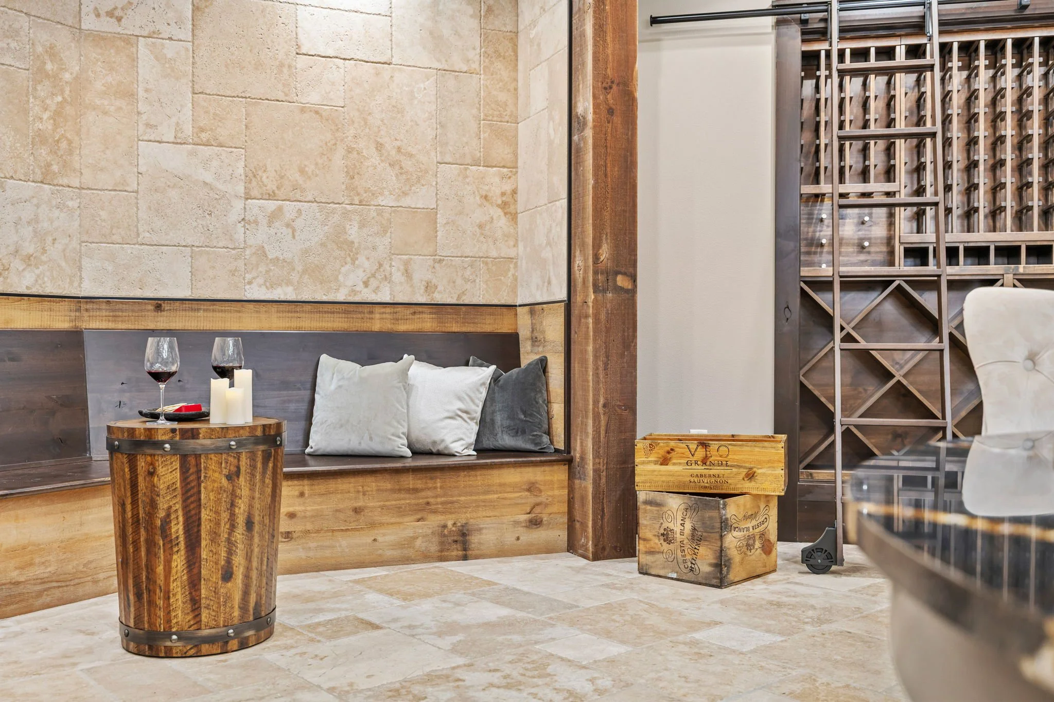 Interior of a room with a wooden bench containing pillows, a small wooden barrel table with two glasses of red wine and candles, a wooden wine crate, and a wine rack in the background.