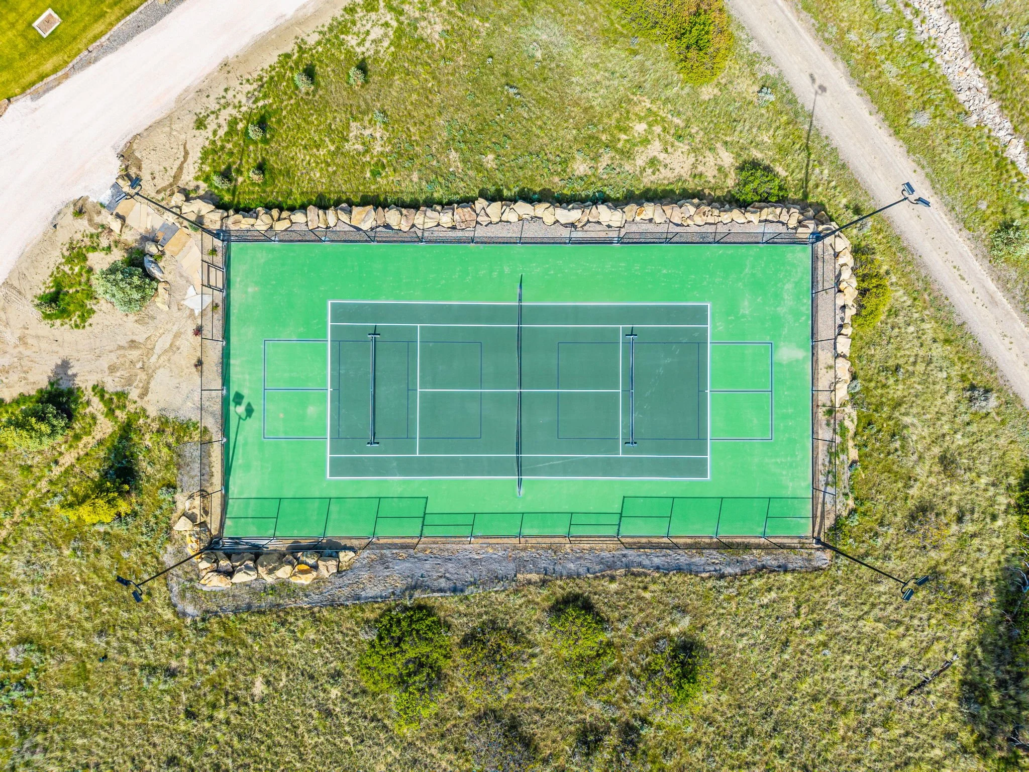 Aerial view of a green tennis court surrounded by a fence and landscaped with rocks and plants.
