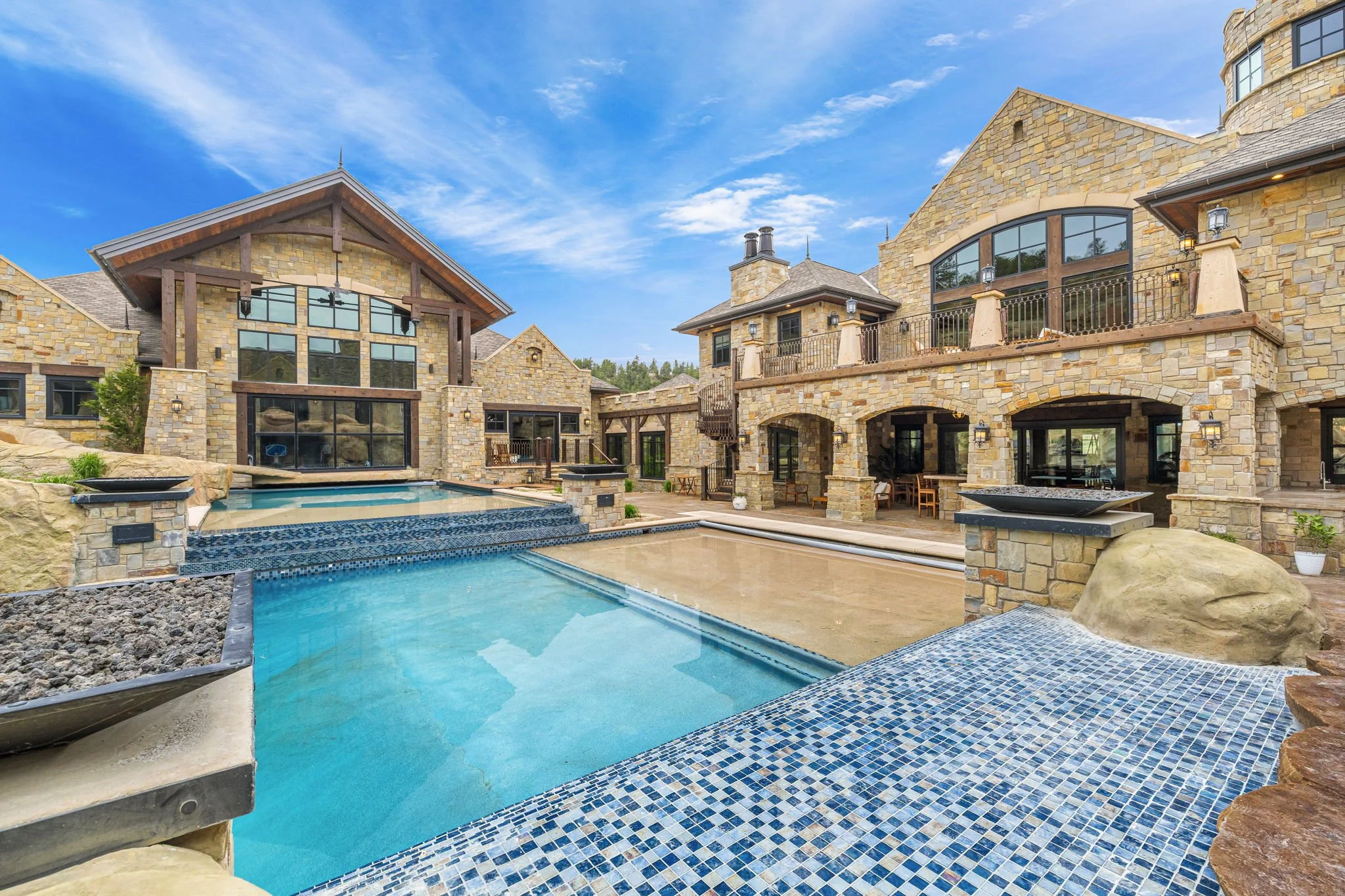 Large luxury house with stone exterior, multiple windows, and balconies, featuring a swimming pool in backyard with clear blue water, mosaic tiles, and patio area under a bright blue sky.