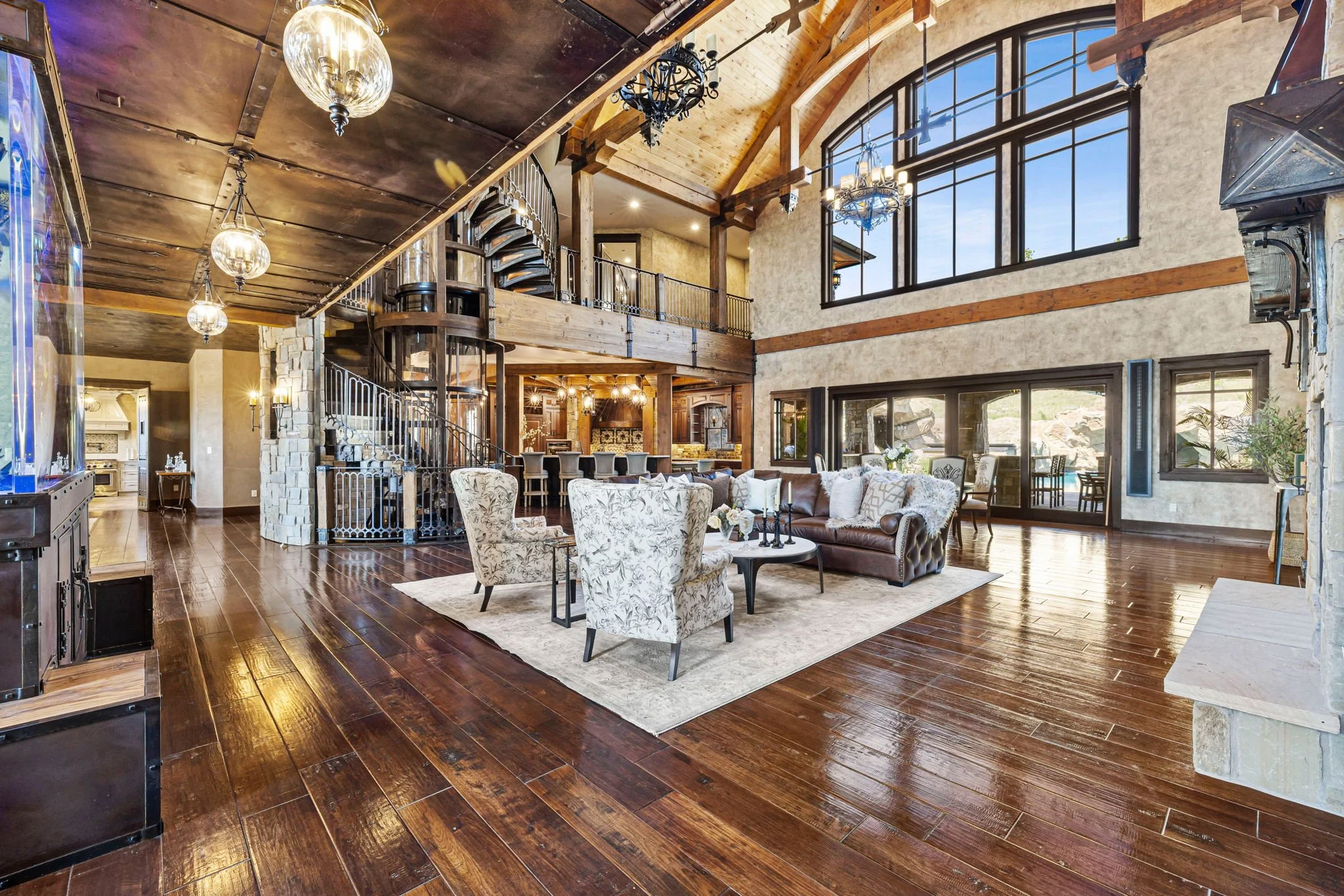 Spacious living room with large windows, wood beams, and a high ceiling. Includes a seating area with a brown leather sofa, patterned armchairs, and a light-colored rug. Decor elements include chandeliers, wall-mounted lights, and modern furnishings,