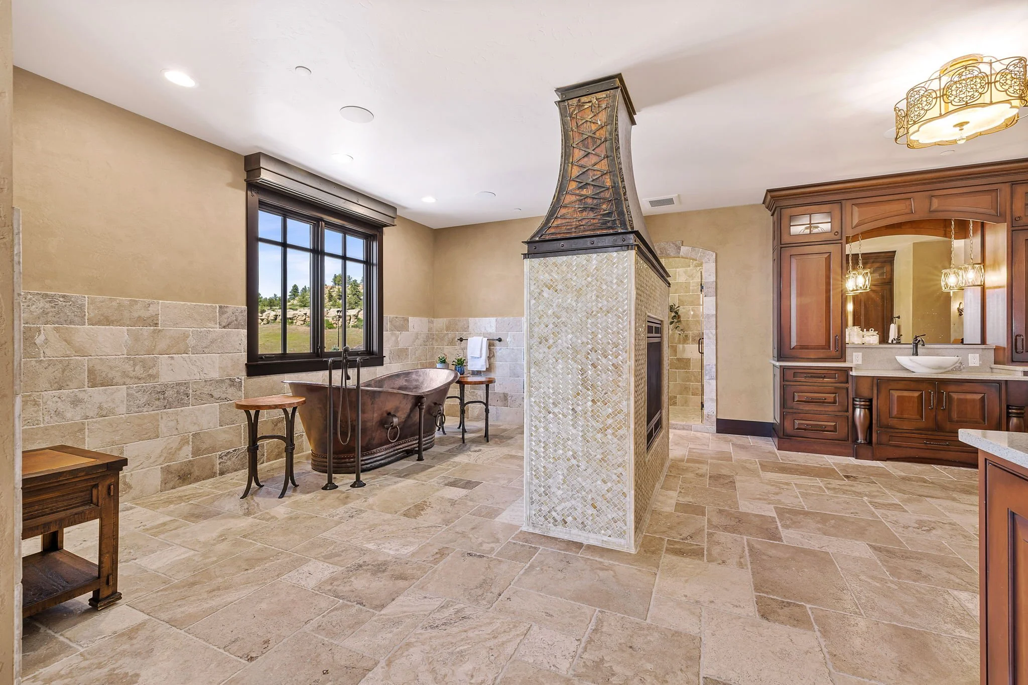 Luxurious bathroom with a freestanding copper bathtub near a window, a tiled central fireplace with a hood, and wooden cabinetry with a mirror and sink.