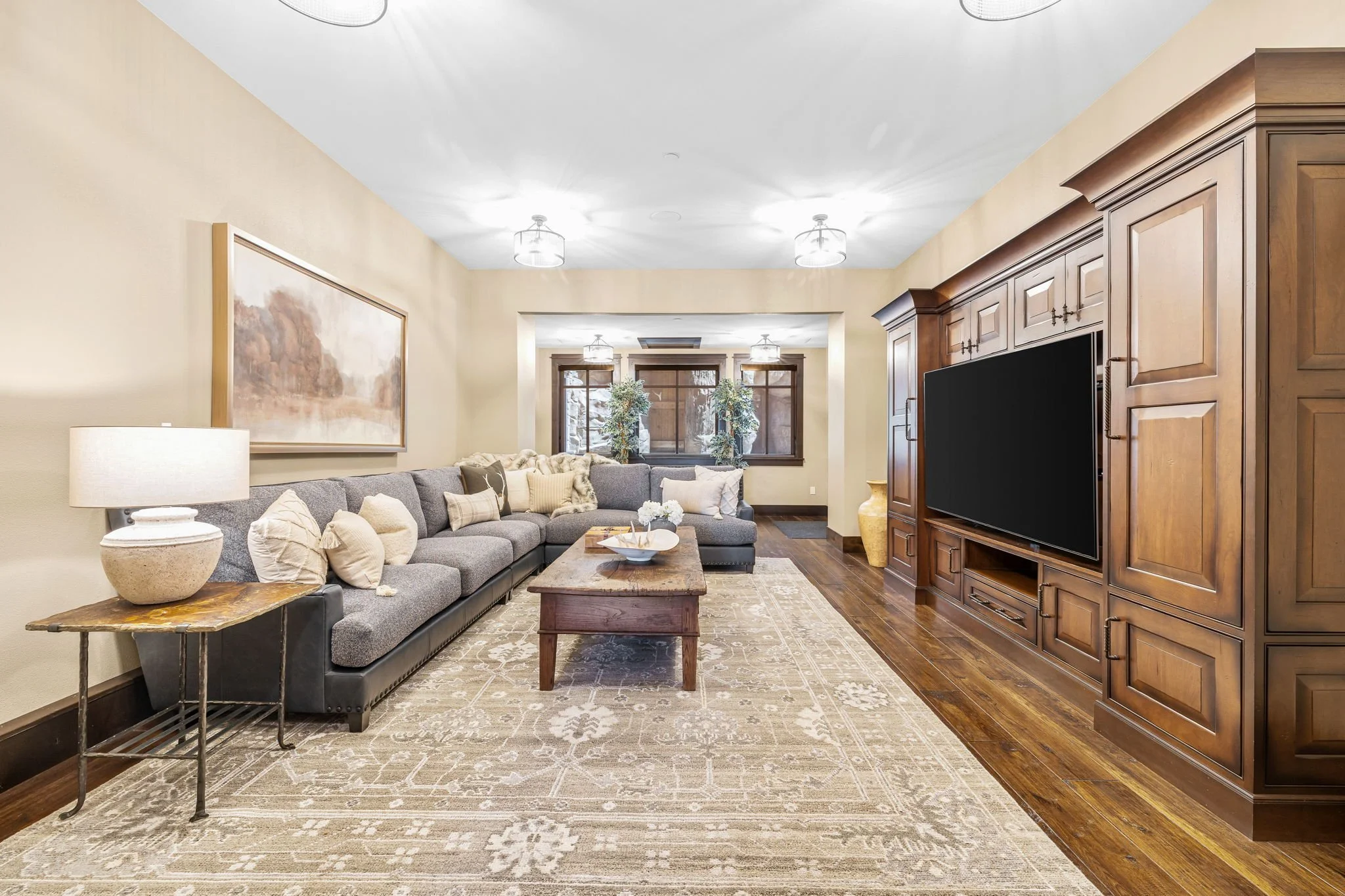 Living room with gray sectional sofa, wooden coffee table, beige area rug, wood entertainment center with a flat-screen TV, side table with a lamp, and large windows with plants.