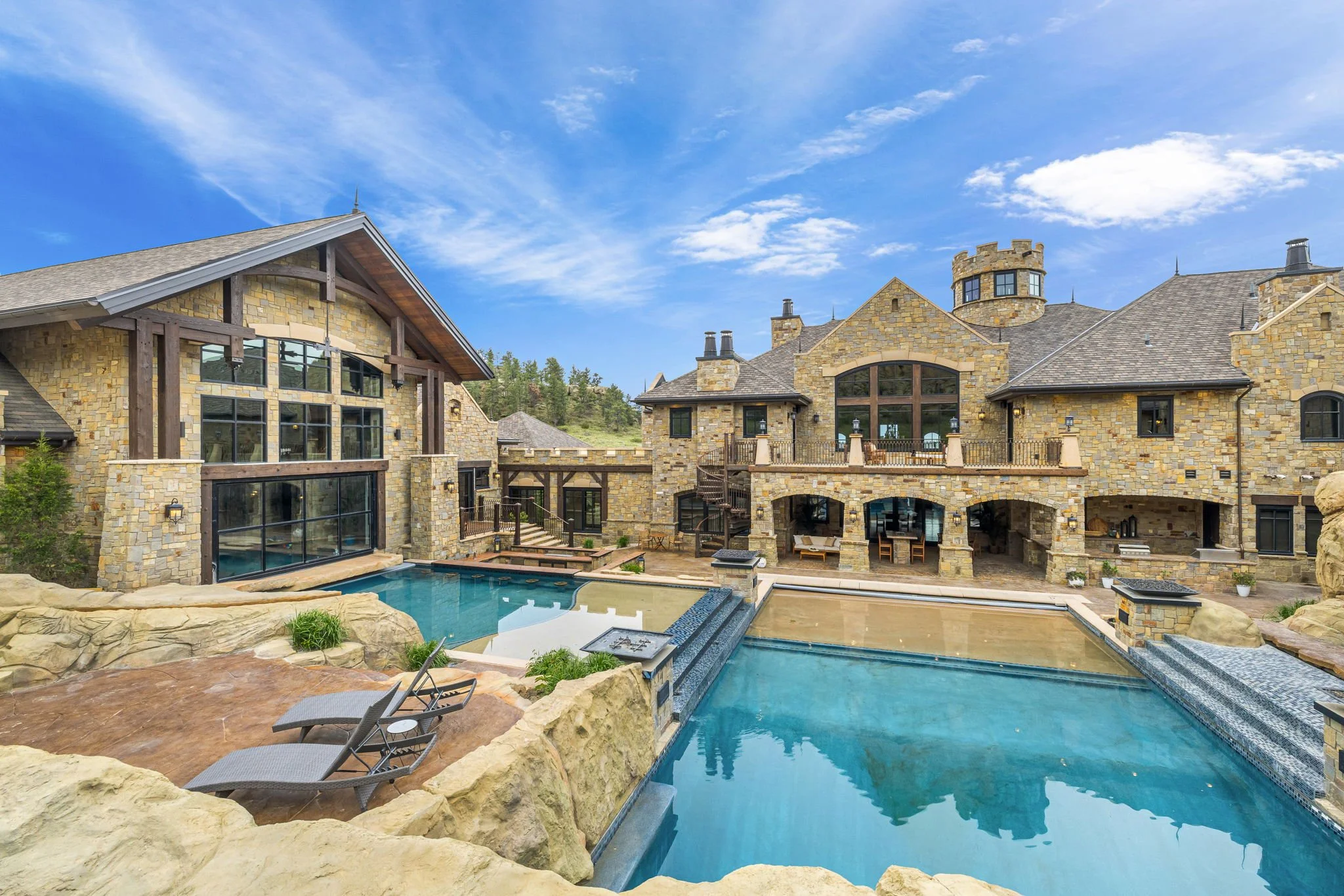 Luxury mansion with a swimming pool and outdoor seating area, stone walls, large windows, and a scenic hillside background under a partly cloudy sky.
