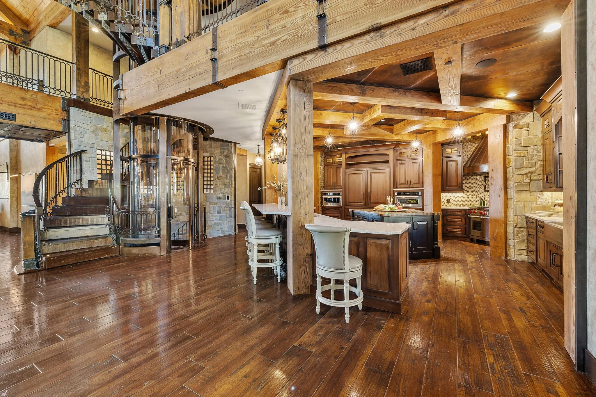 Interior view of a rustic kitchen with wooden beams, dark wood cabinetry, stone wall accents, hardwood floors, and a spiral staircase with glass enclosure.