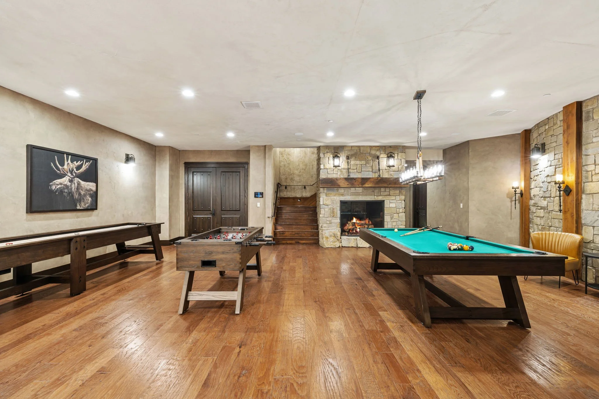 Interior of a game room with a wood fireplace, two pool tables, a foosball table, wooden stairs, a framed moose photo, wooden flooring, and warm lighting.