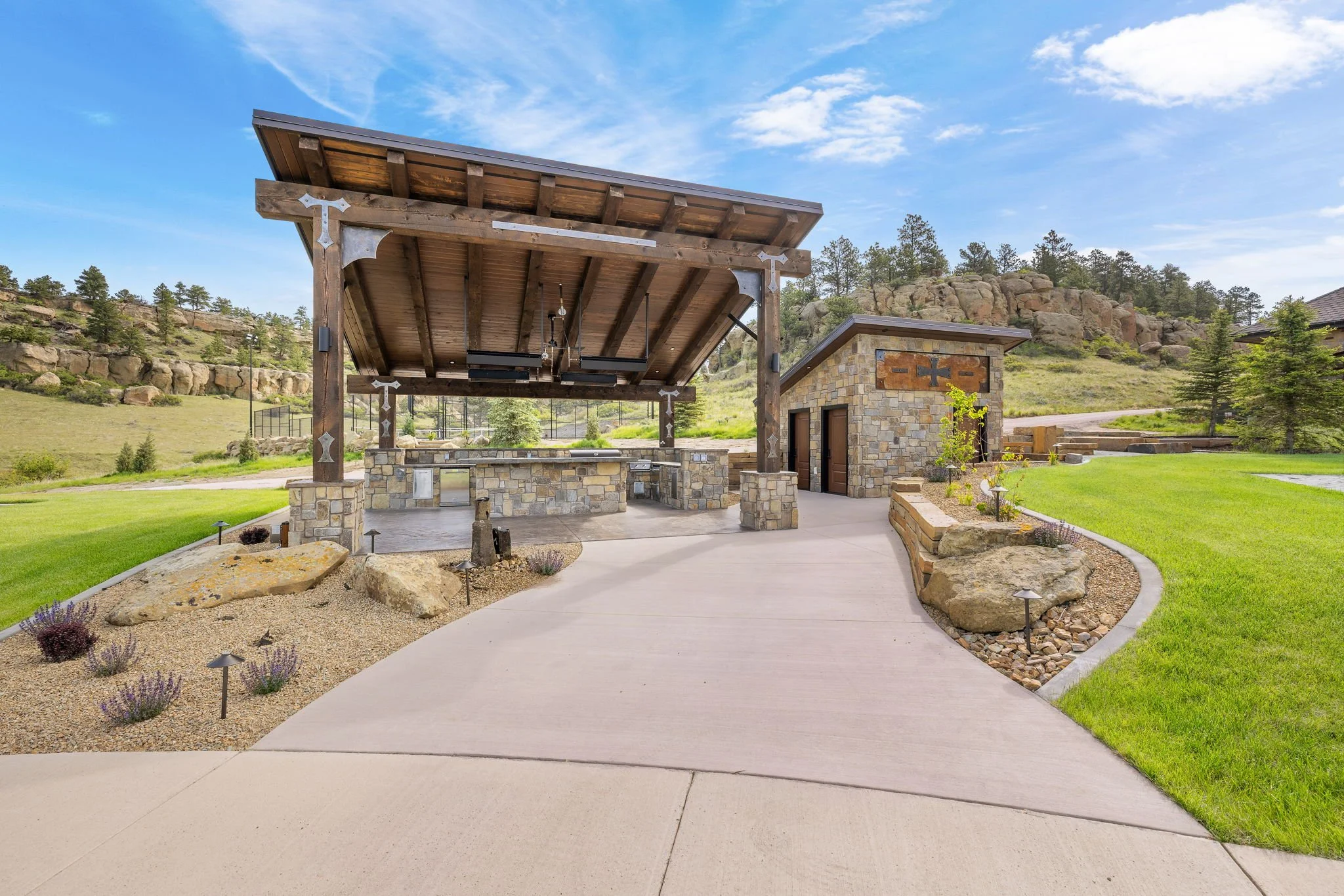 An outdoor stone and wood pavilion with a barbecue area, set in a landscaped yard with rocks, plants, and a grassy lawn, surrounded by hills and trees.