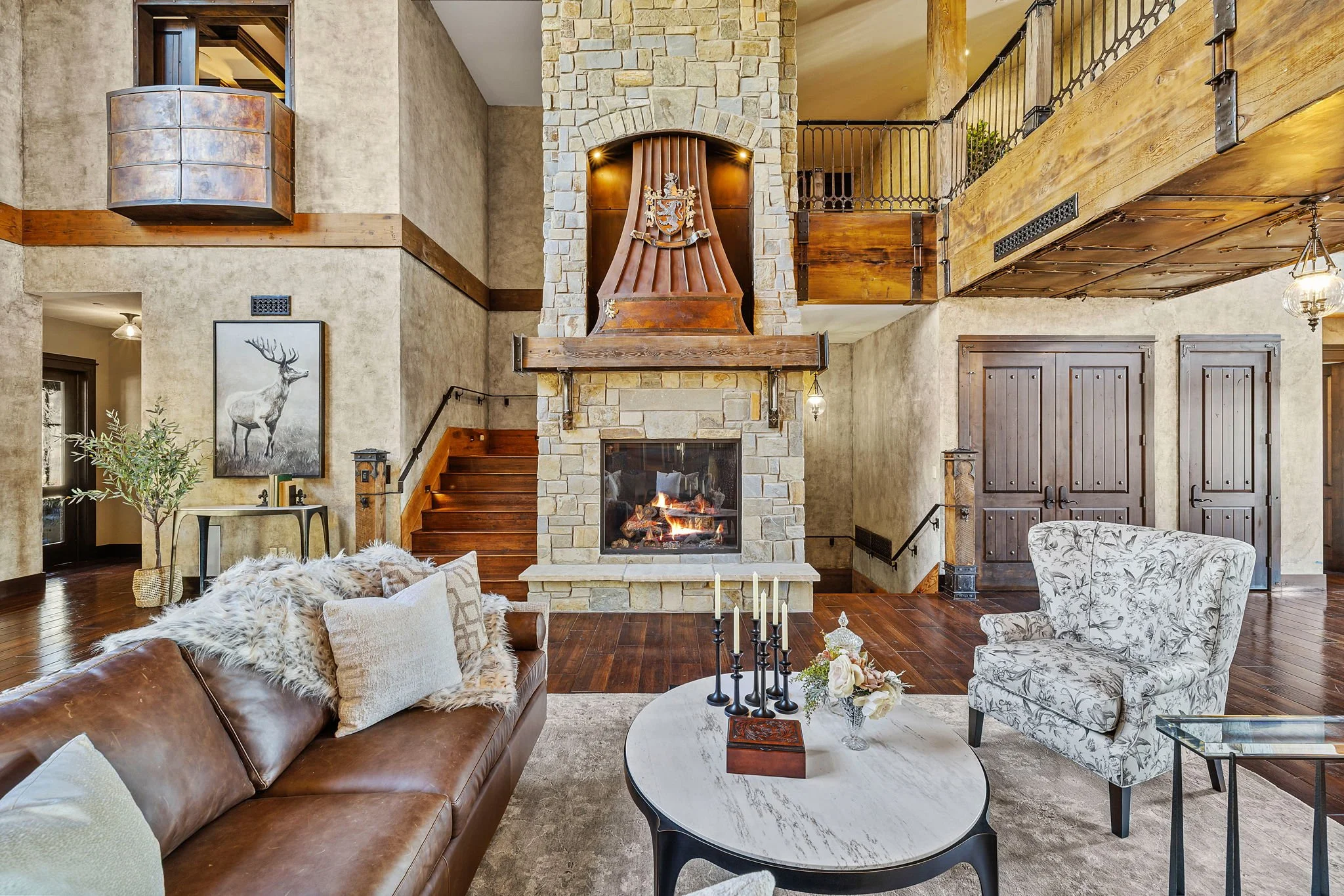 A cozy living room with a stone fireplace, wooden accents, and modern furniture. There is a brown leather sofa with pillows and a fluffy throw, a patterned armchair, and a marble-top coffee table with candles and floral decor. The room has an open la