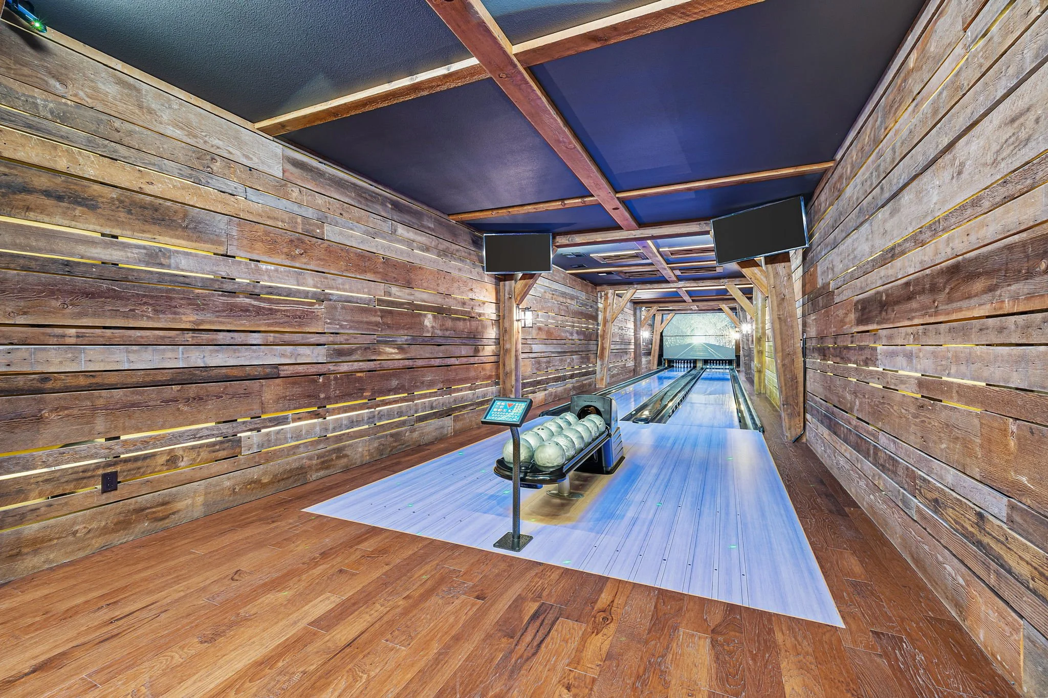 Indoor bowling alley with wooden wall panels, two large TV screens, and a bowling ball return system with ten bowling balls on it. The floor is a light wood color with a blue bowling lane extending towards the pins.
