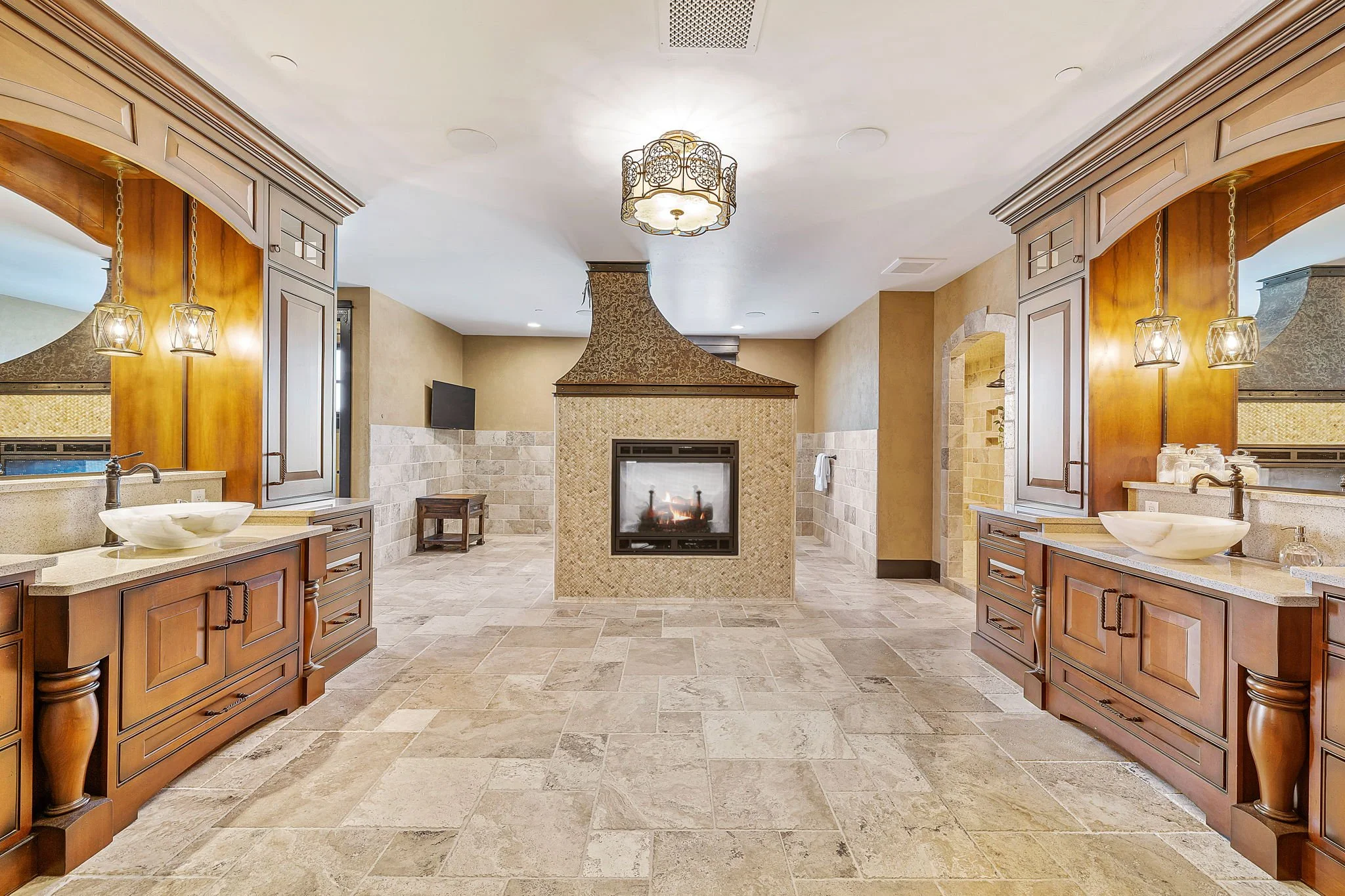 A spacious, well-lit bathroom with two wooden vanities, each with a vessel sink and decorative lighting, a central fireplace, beige tiled flooring, and a small seating area in the back.