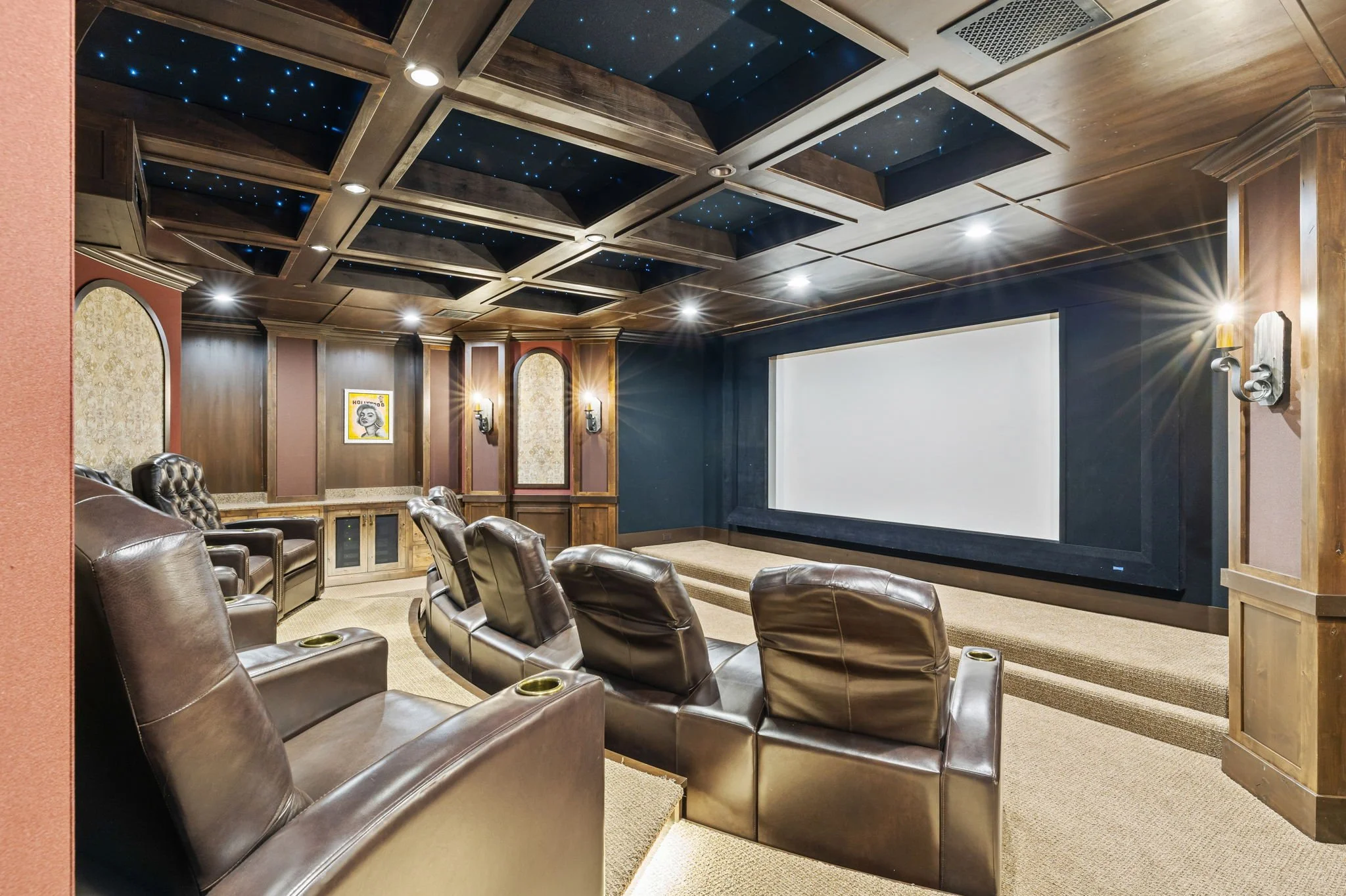 Luxury home theater room with brown leather recliners, a large projection screen, wooden paneling, decorative wall sconces, a patterned wallpaper accent wall, and a ceiling with black panels decorated with blue star-like lights.