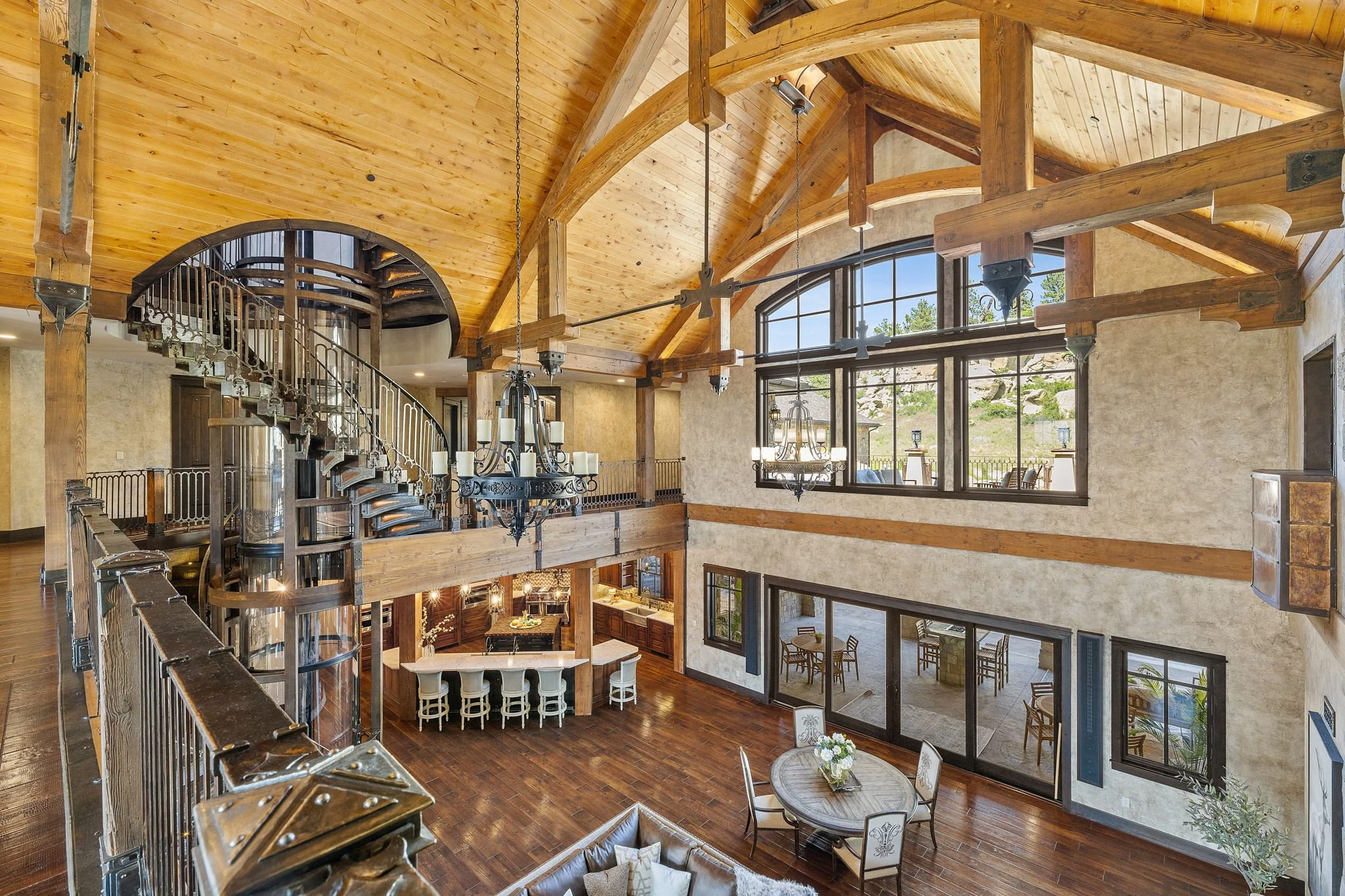 A spacious living area with a high, vaulted wooden ceiling, large windows, hardwood floors, and a mix of modern and rustic decor. There is a curved staircase, a chandelier, a dining table with chairs, and an open kitchen area.