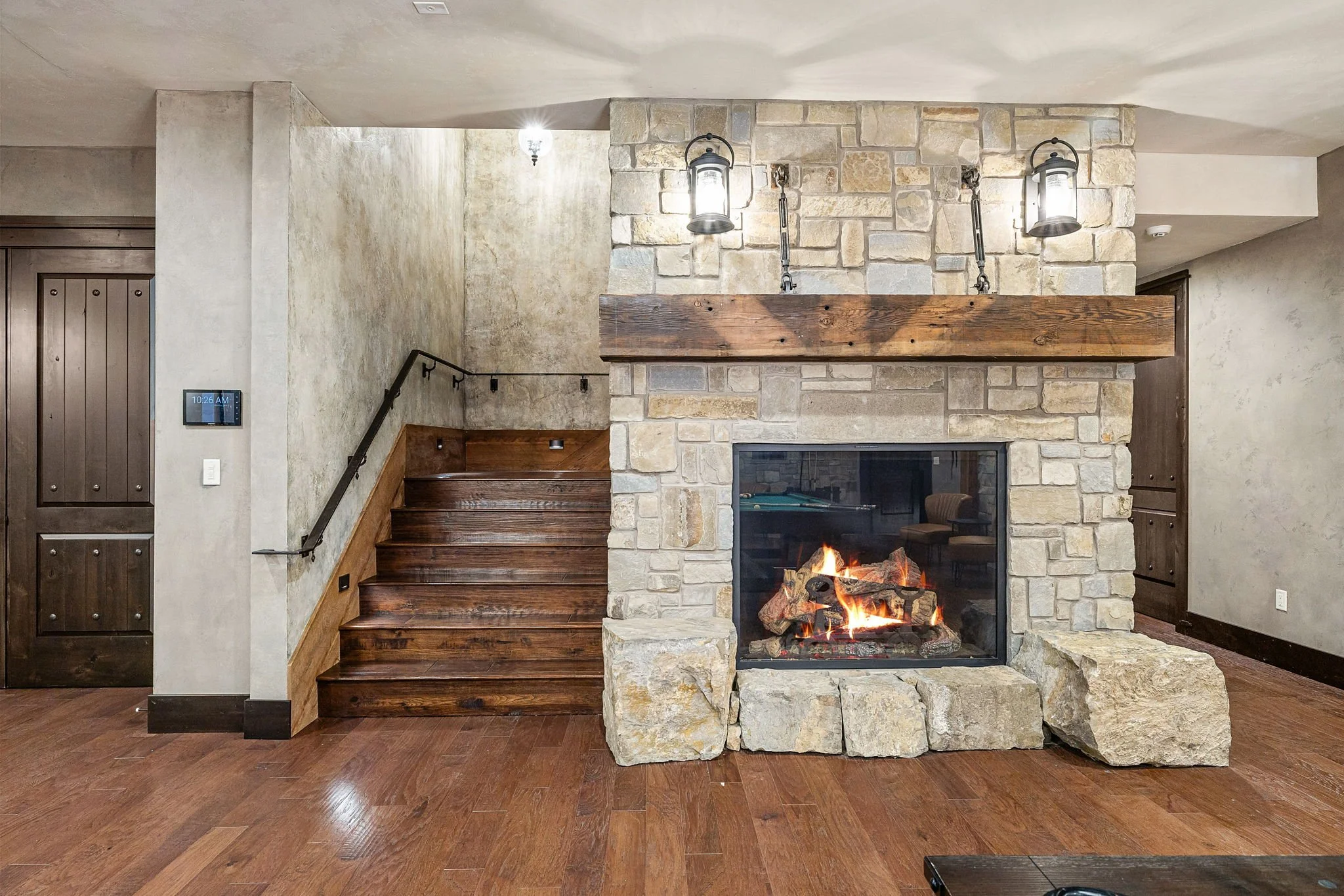 Living room with stone fireplace, wooden mantel, and wooden stairs, illuminated by two wall-mounted lantern-style lights.